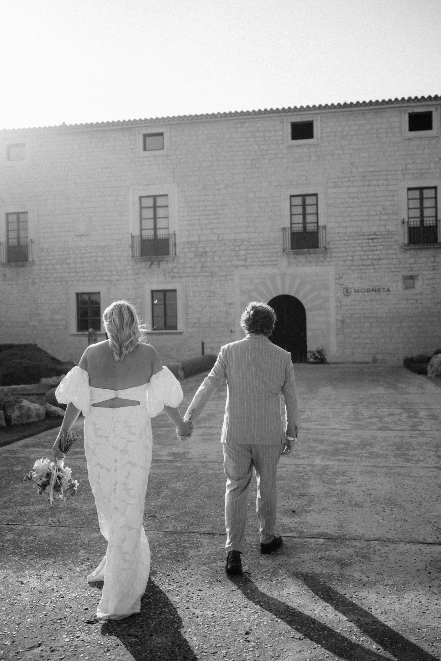 Black and white photo of bride and groom walking hand in hand toward a historic stone wedding venue.