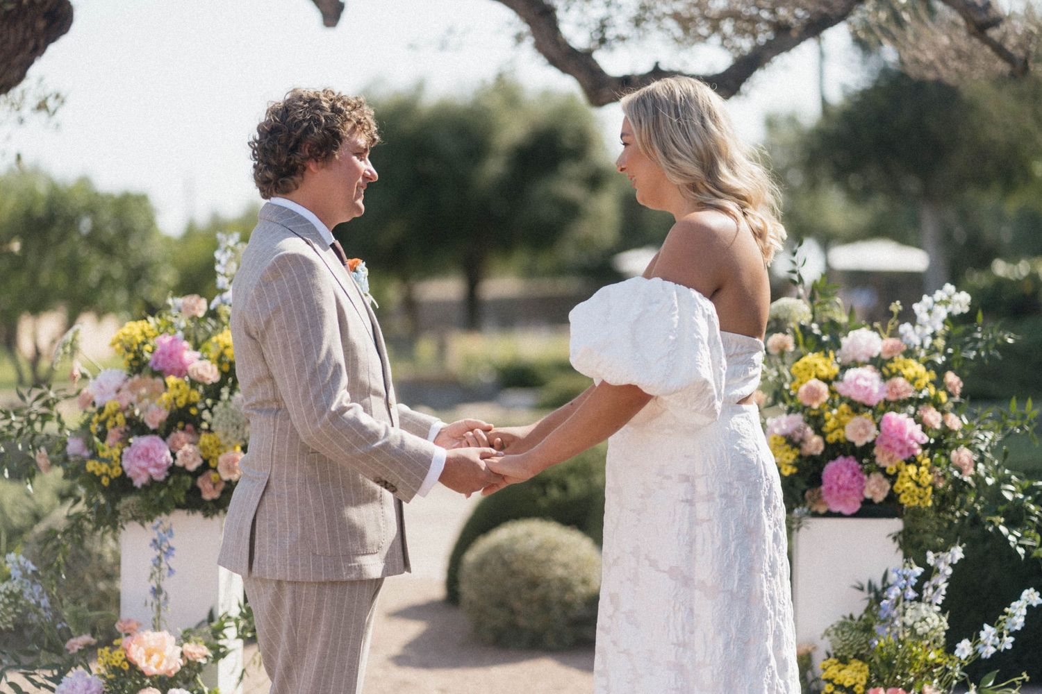 Couple exchanging vows at an outdoor wedding ceremony surrounded by colorful pink and yellow floral arrangements.