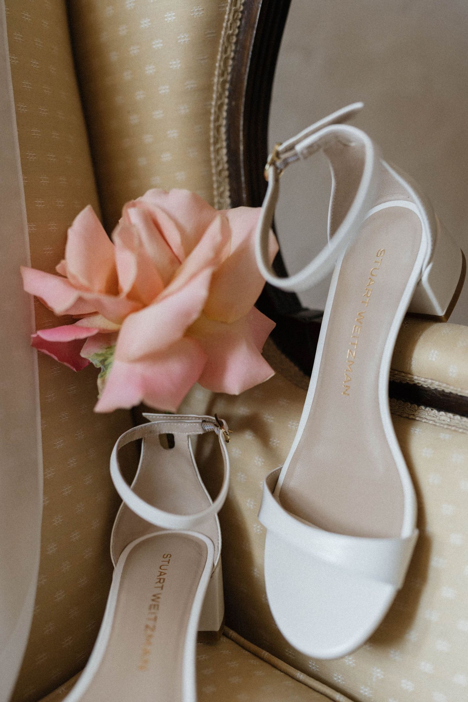 White bridal heeled sandals by Stuart Weitzman displayed on a chair beside a soft pink rose.