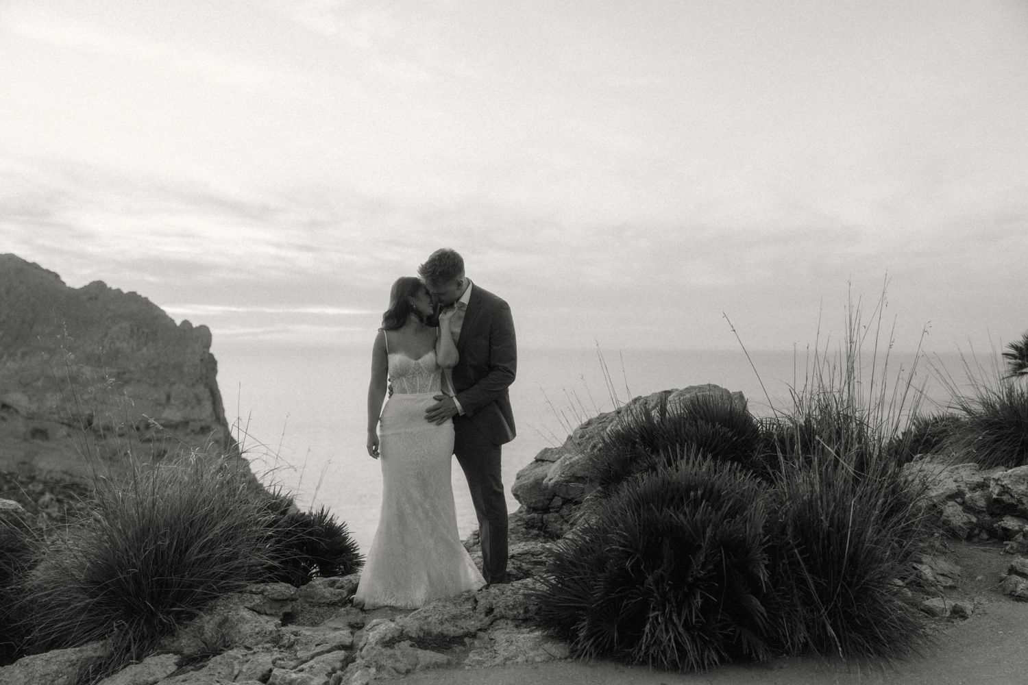 Black and white photo of bride and groom sharing a tender kiss on coastal cliffs with ocean view at dusk.