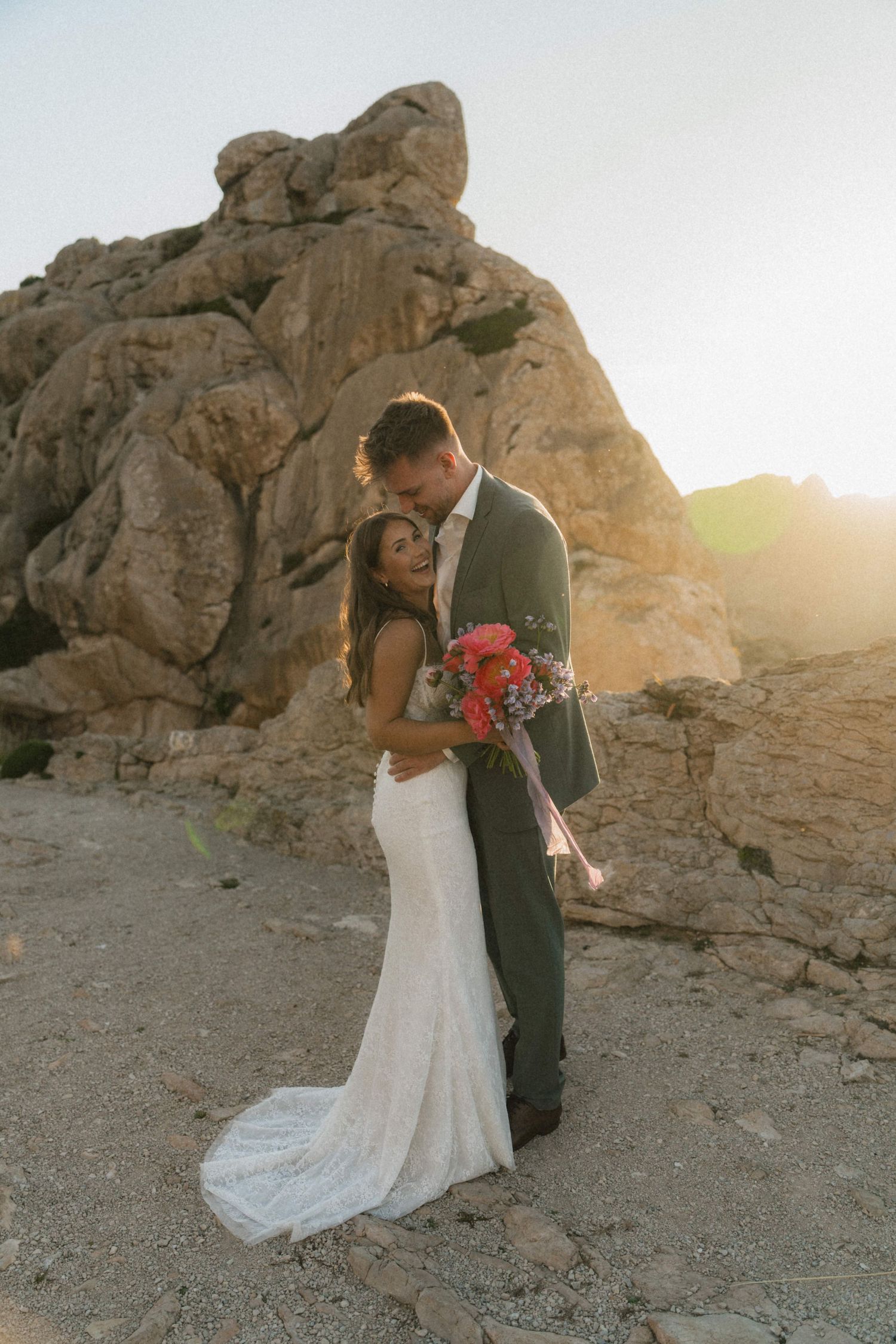 Bride and groom embrace at golden hour beside dramatic rocky formations in a desert landscape.