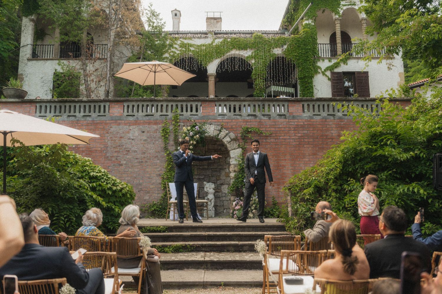 Two grooms standing at outdoor wedding altar in historic villa garden with seated guests.
