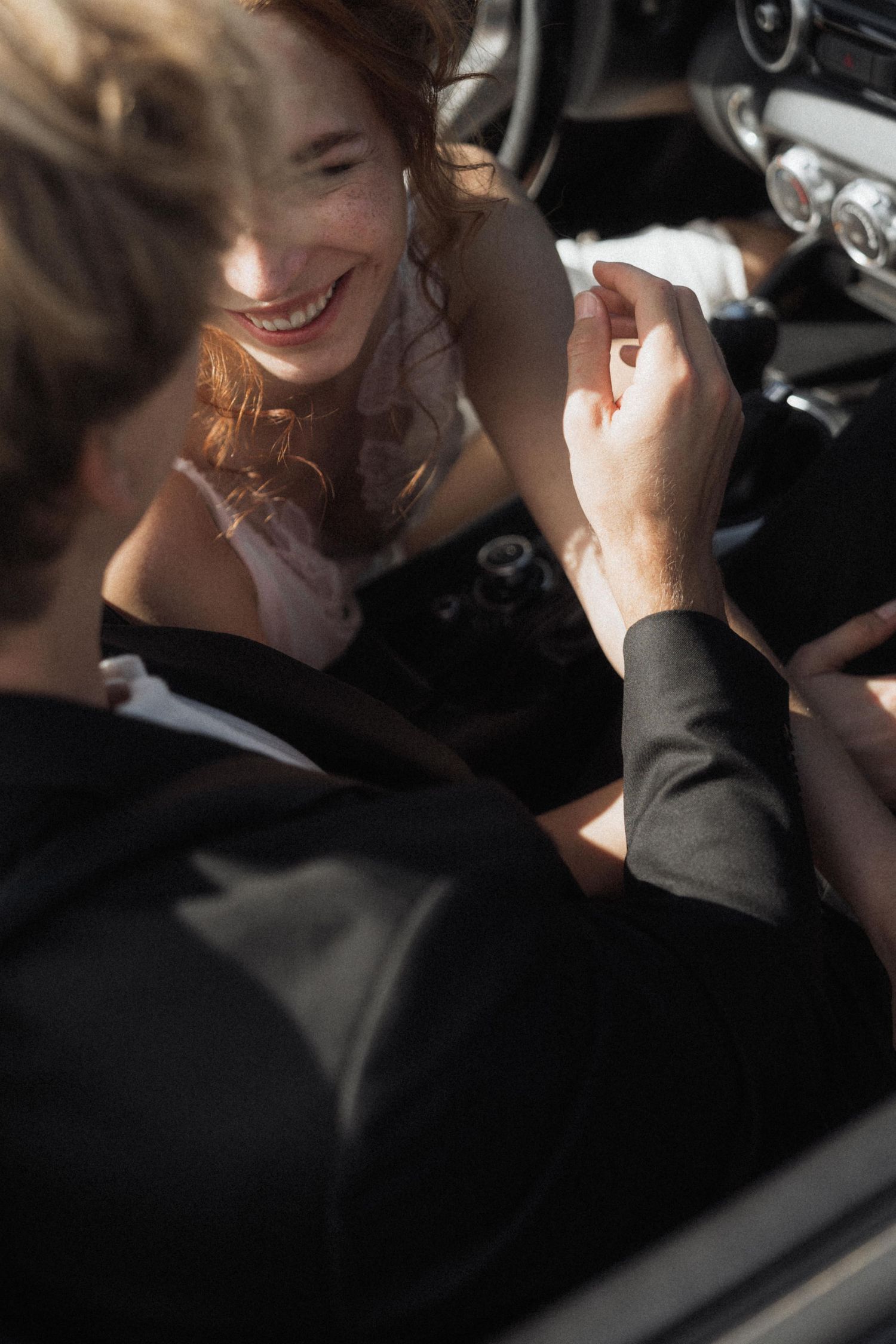 Smiling redhead bride holding hands with groom in black suit inside a vintage car on wedding day.