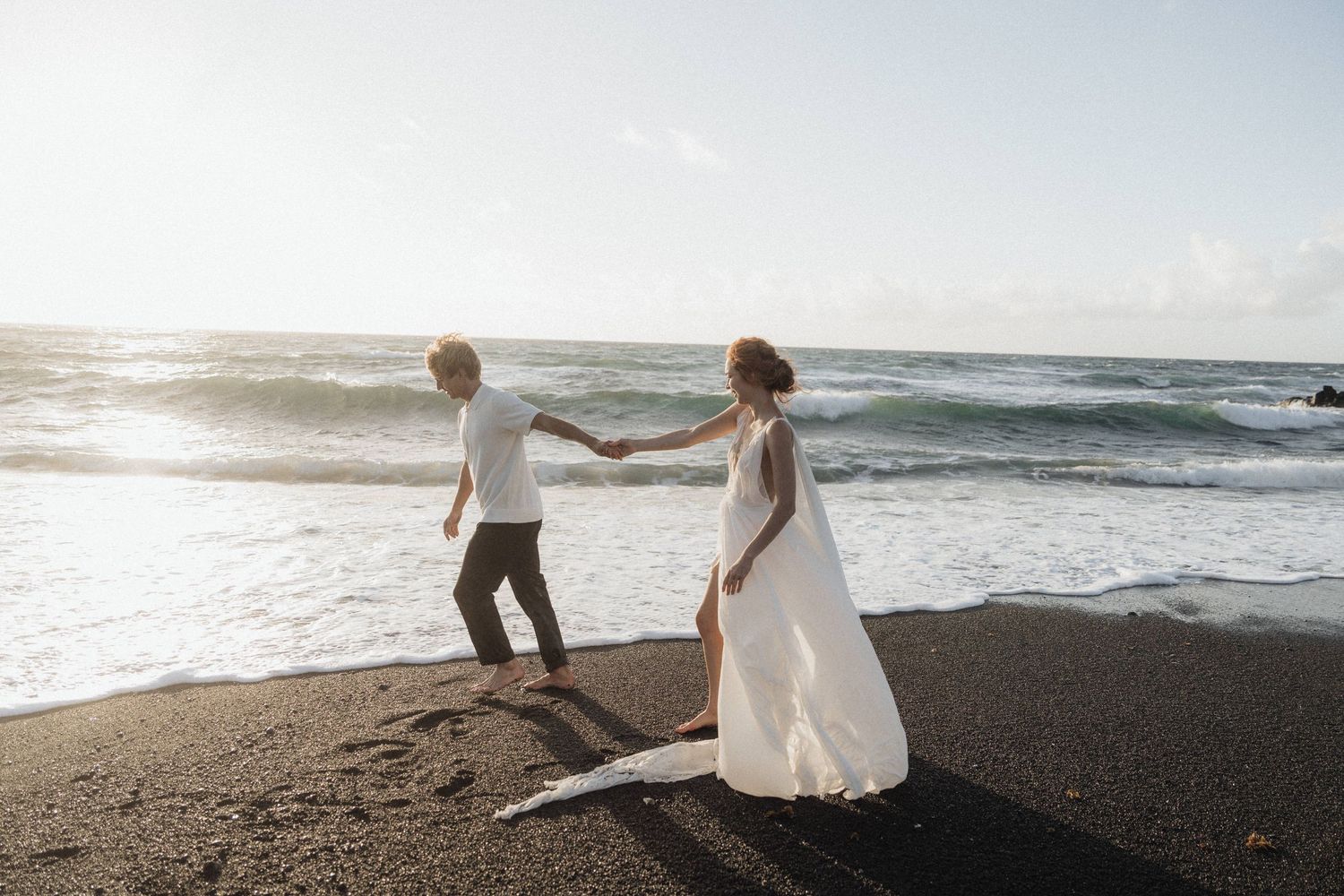 Bride and groom holding hands walking along a black sand beach at sunset.