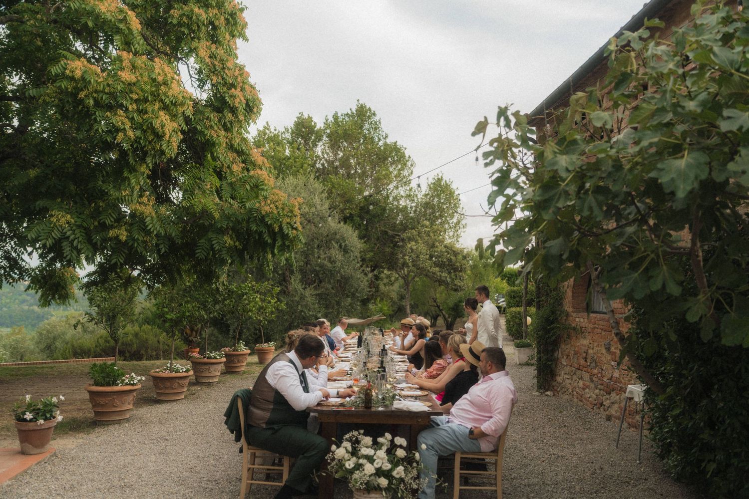 Guests dining at a long outdoor table beside a rustic stone building surrounded by lush greenery.