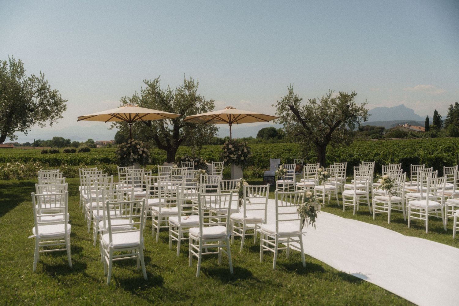 Elegant outdoor wedding ceremony setup with white Chiavari chairs, a white aisle, and patio umbrellas amid lush greenery.