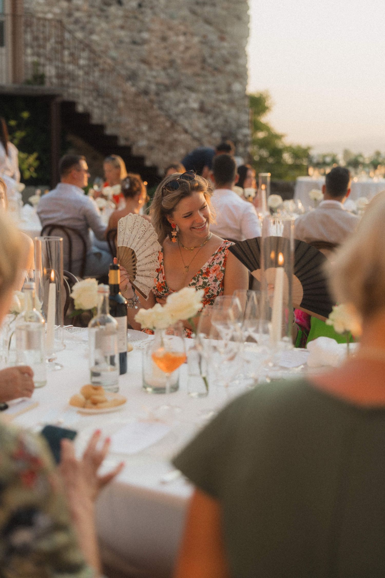 Wedding guest smiling with hand fan at elegant outdoor reception dinner table at sunset.