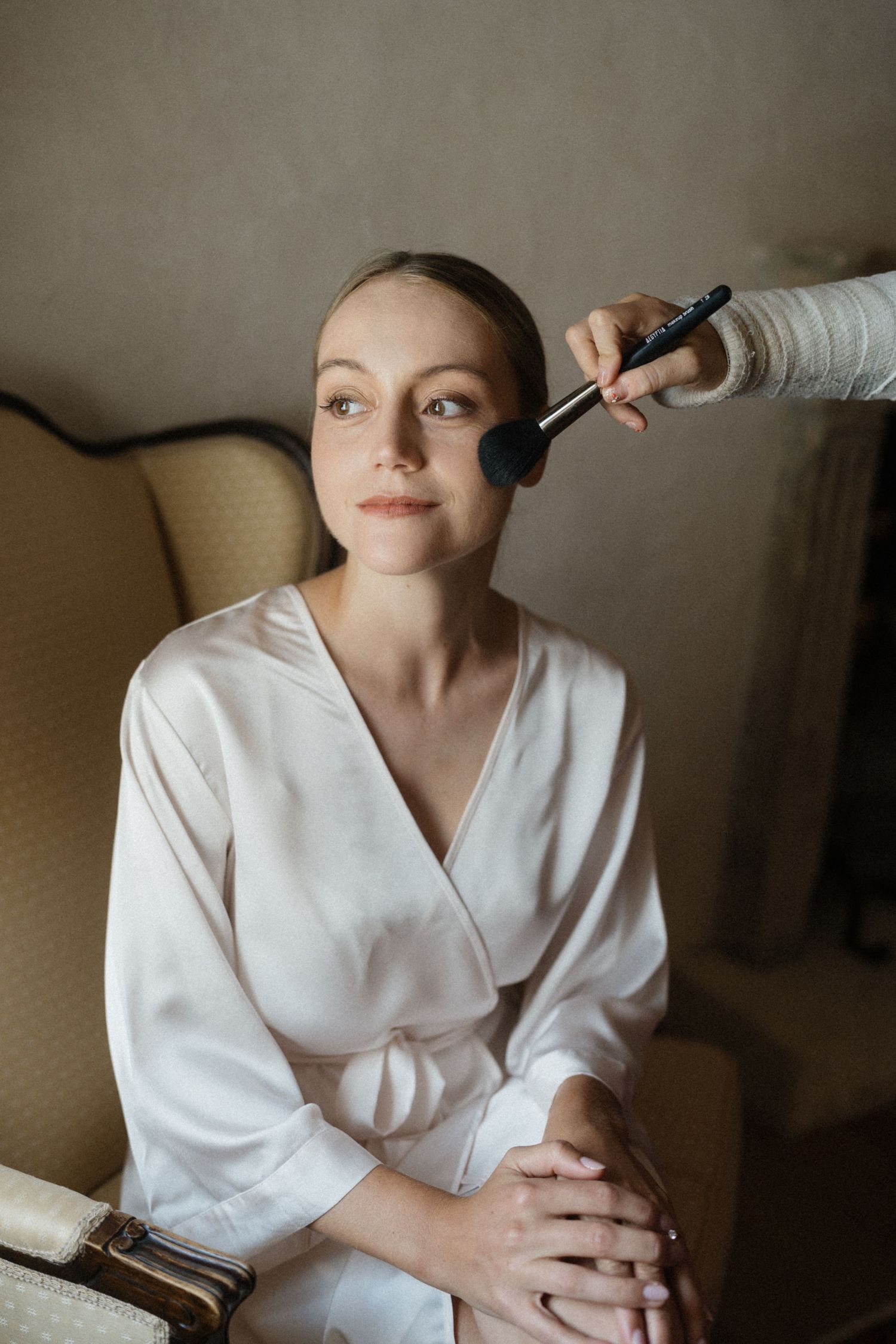 Bride in white silk robe getting professional makeup applied with brush while seated in elegant chair.