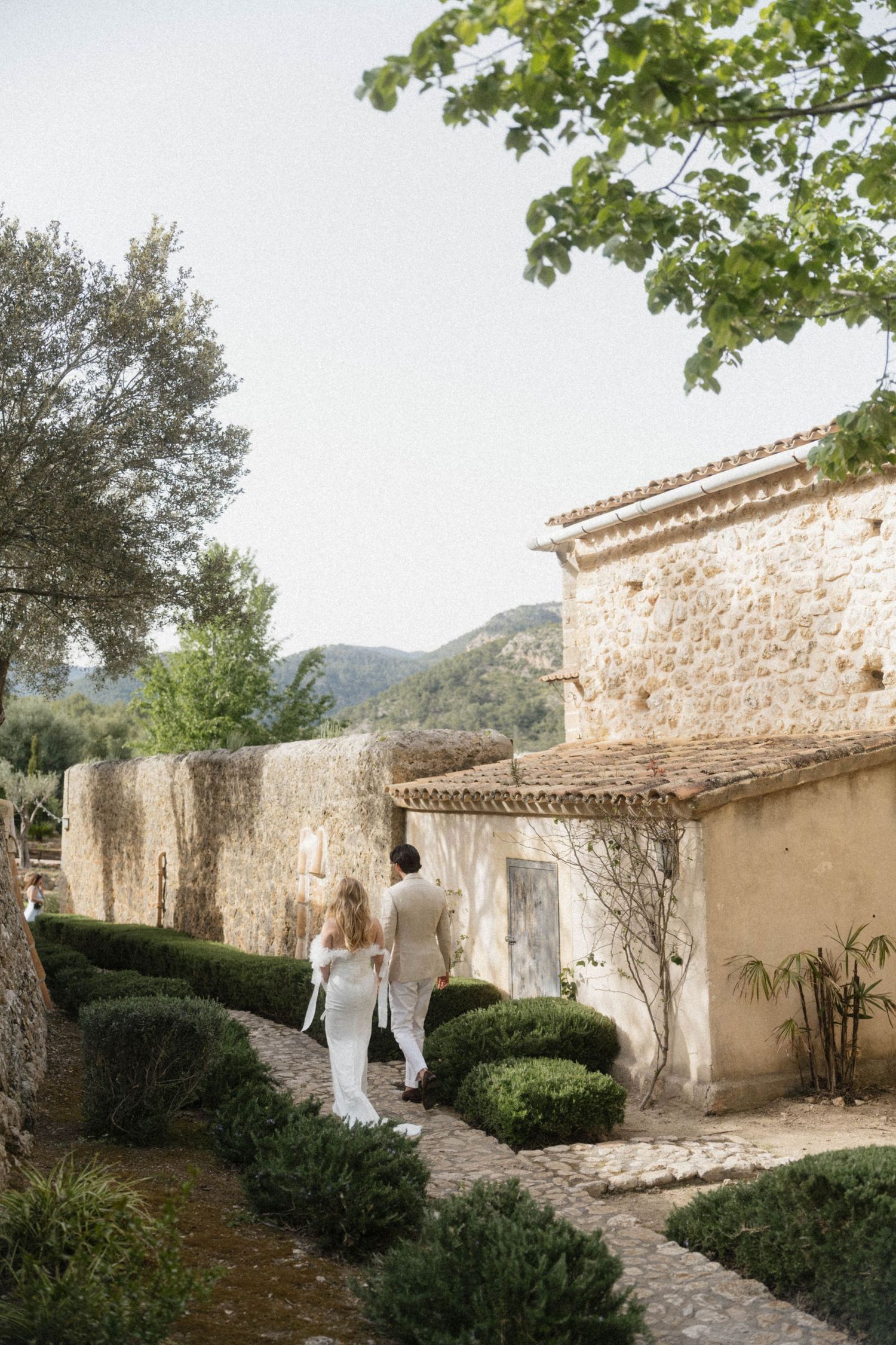 Bride and groom walking through a rustic stone estate garden with manicured hedges and mountain views.