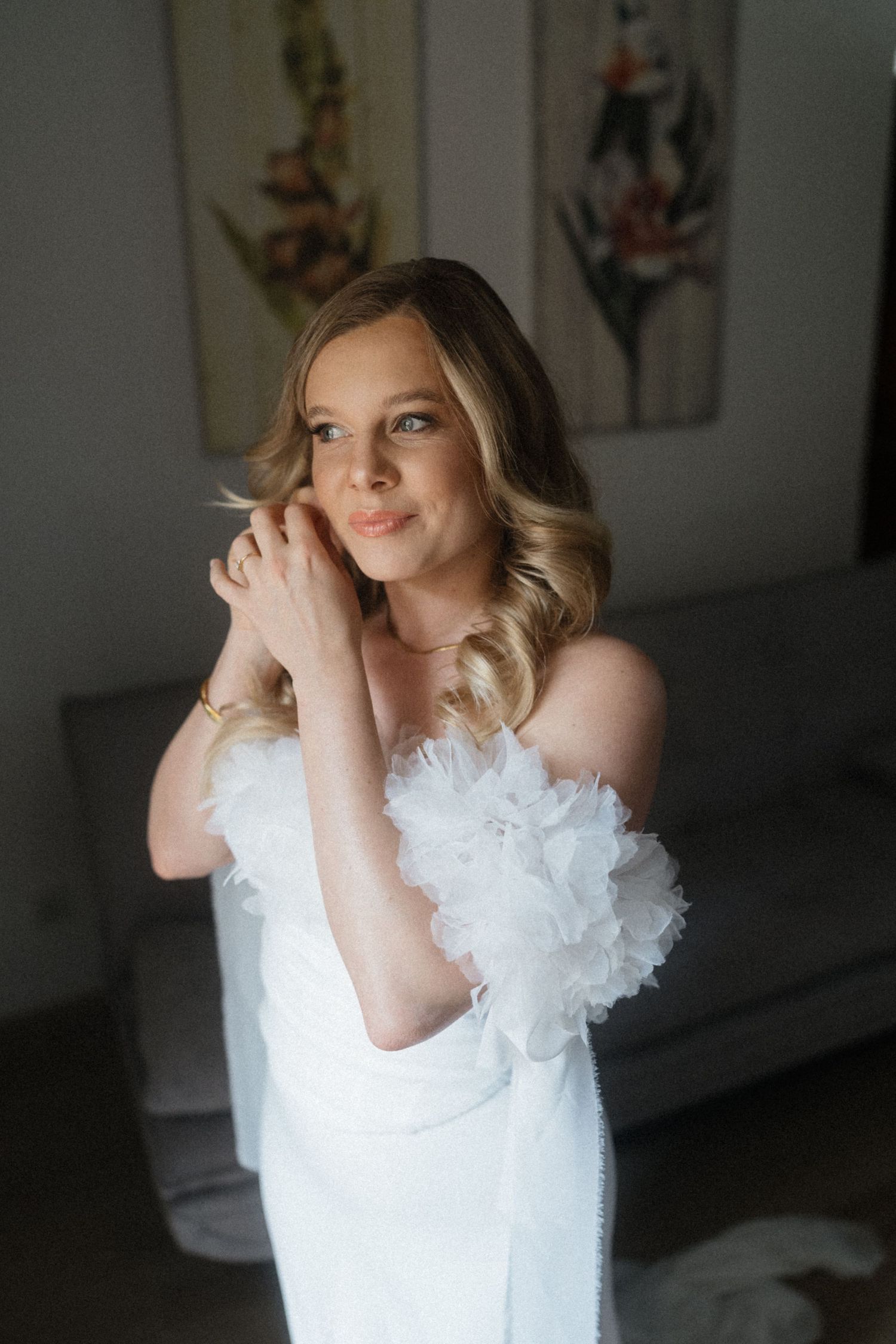 Blonde bride in a white off-shoulder gown with feather details adjusting her earrings before her wedding ceremony.