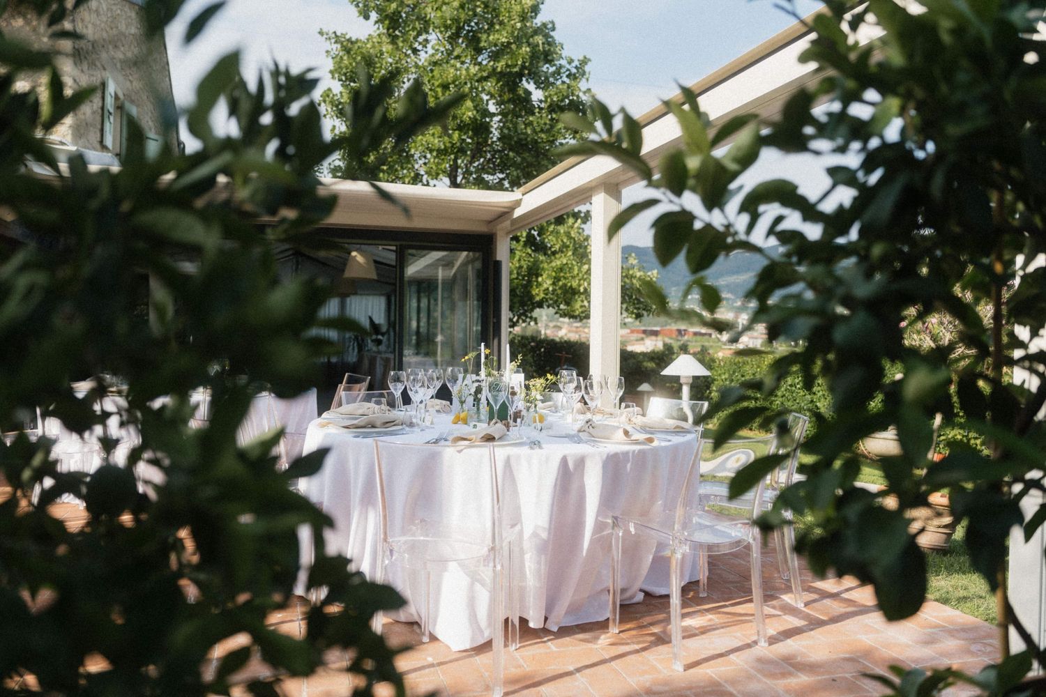Elegant outdoor wedding reception table set with white linens and glassware on a sunlit terrace surrounded by lush greenery.