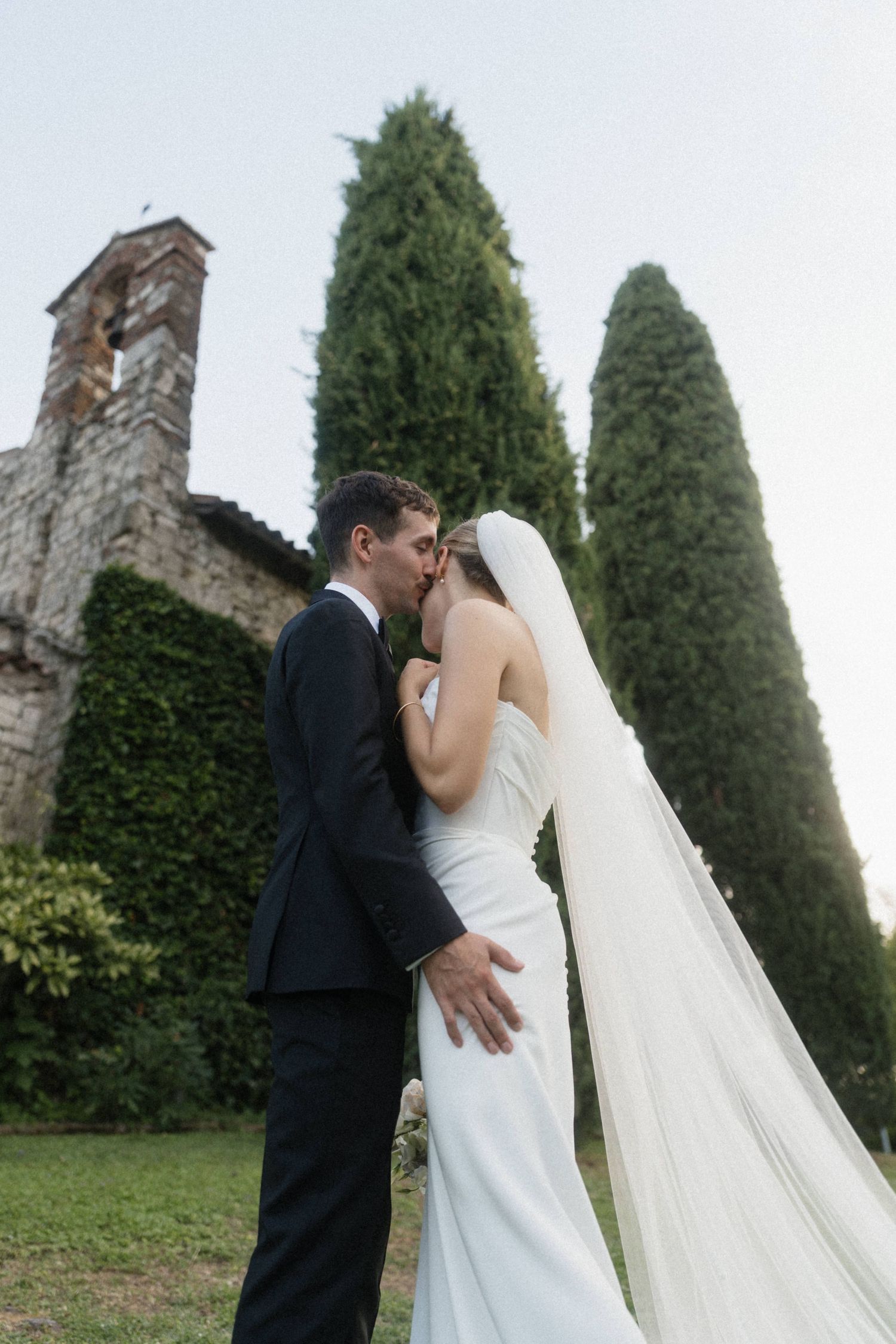 Bride and groom kissing in front of a historic stone church with cypress trees.
