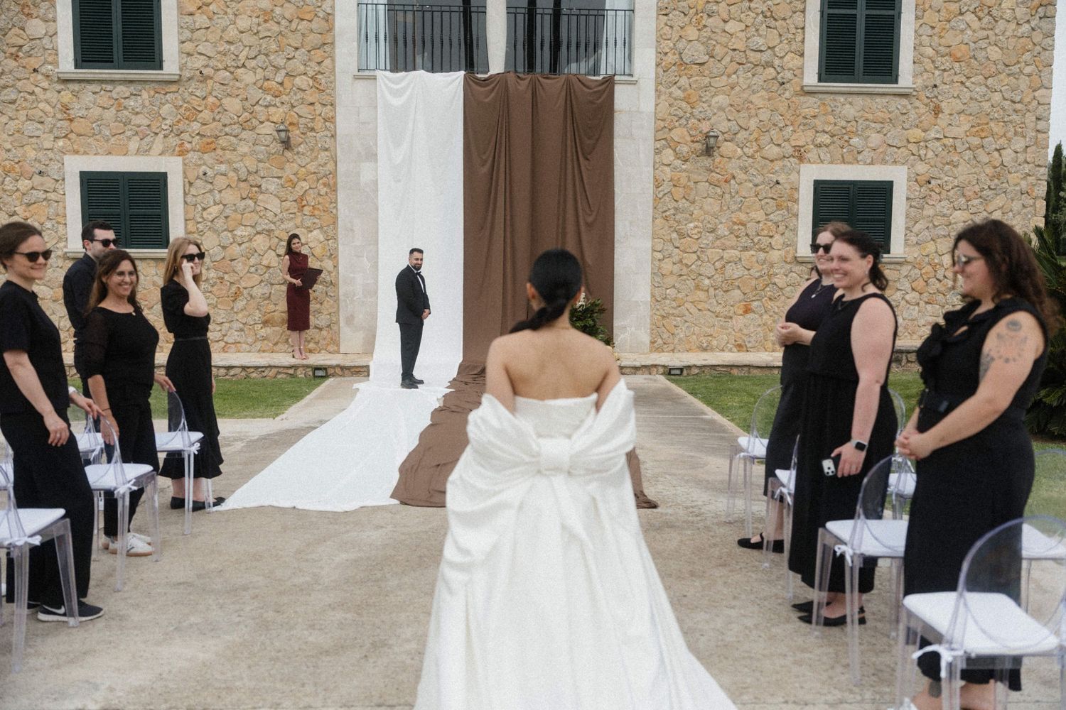 Bride in white gown walks down outdoor aisle toward groom, flanked by bridesmaids in black at elegant stone venue.