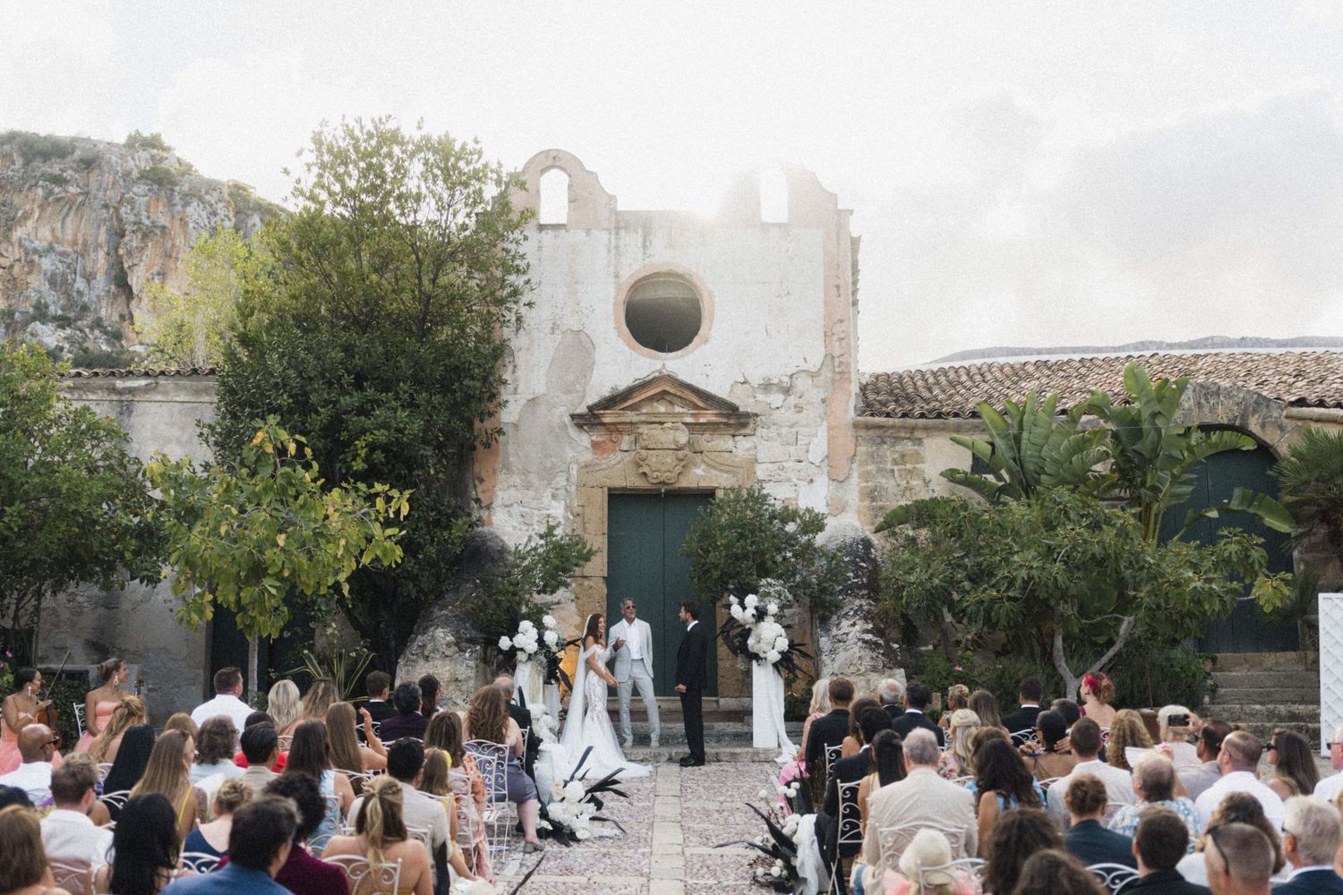 Outdoor wedding ceremony at rustic stone chapel with guests gathered on cobblestone courtyard.