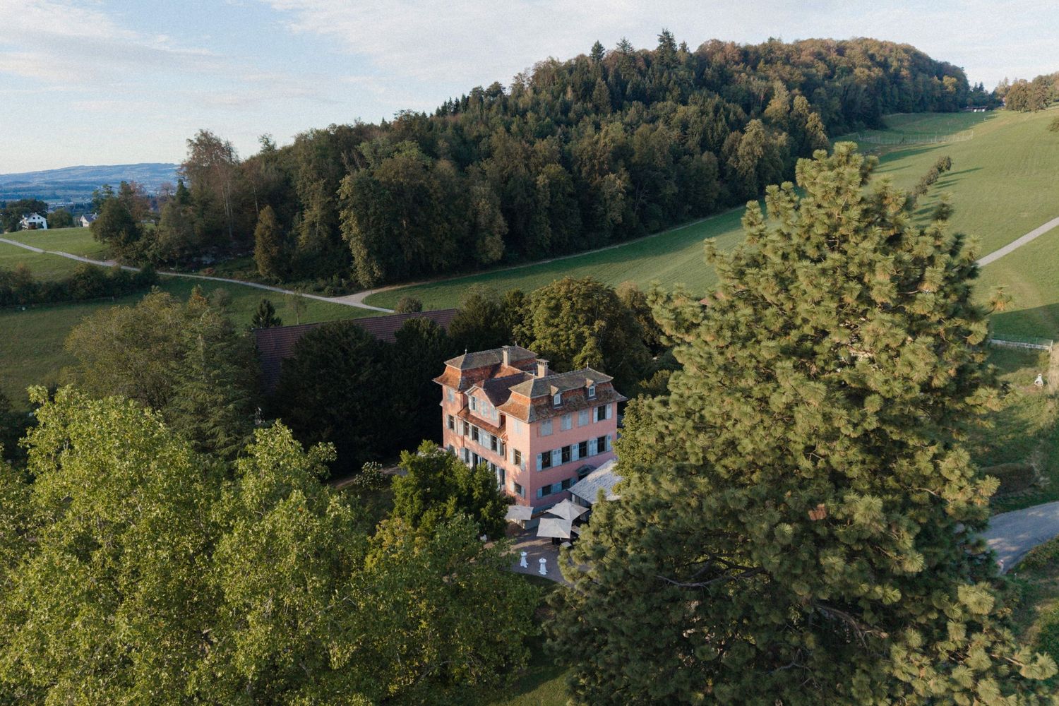 Aerial view of a charming pink villa nestled among lush green trees and rolling hills at golden hour.
