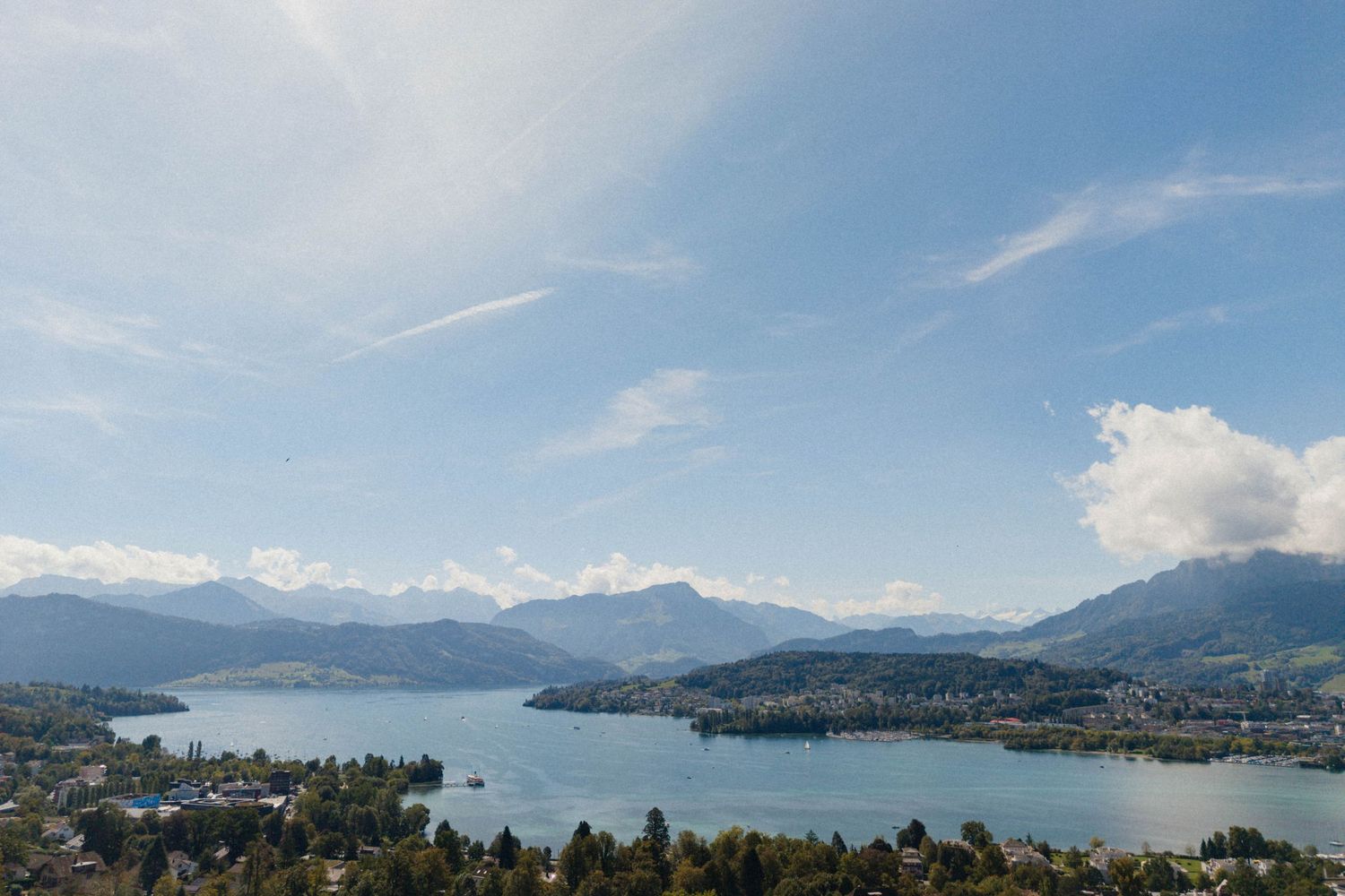 Panoramic view of a serene alpine lake surrounded by lush trees and mountains under a bright blue sky.