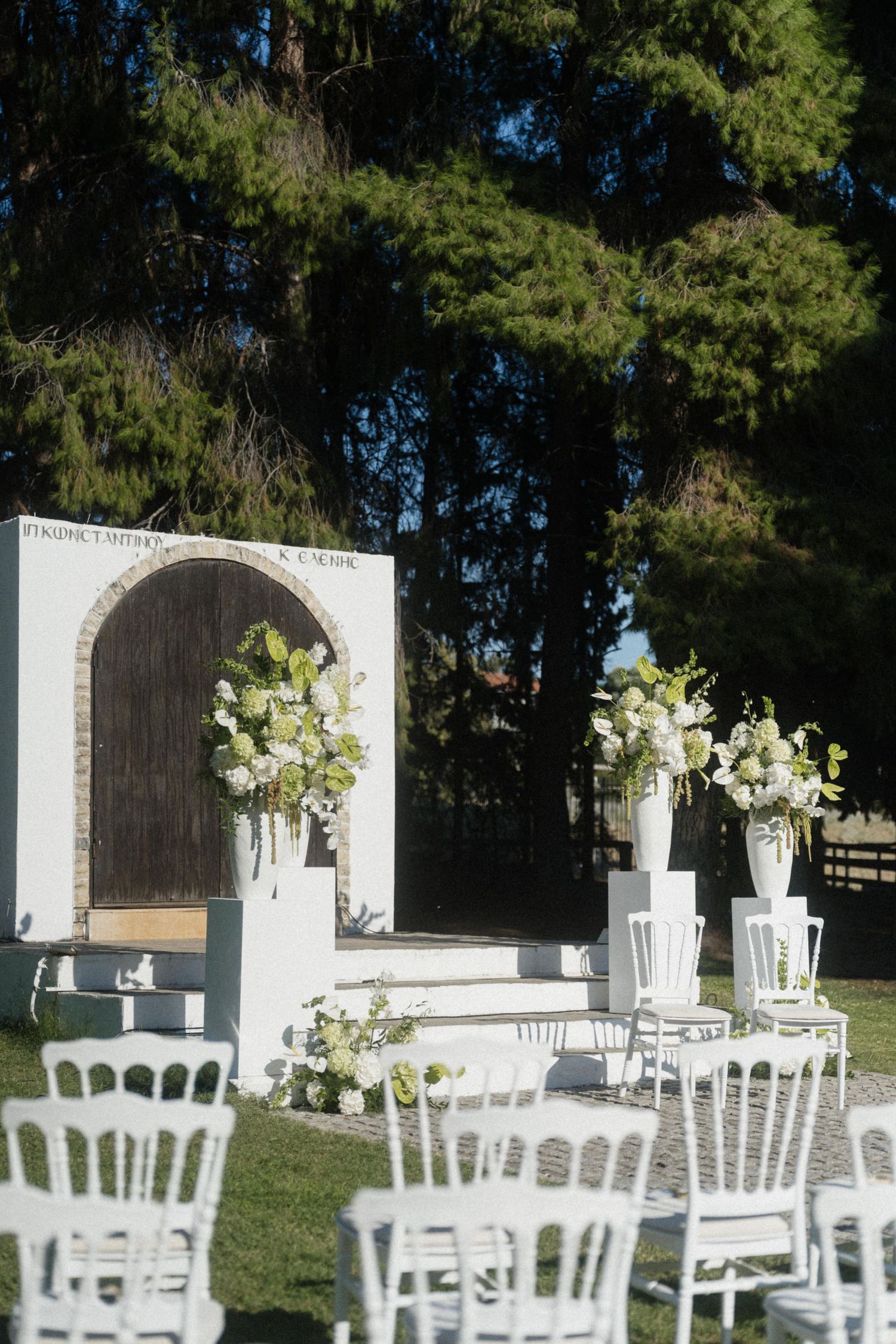 Elegant outdoor wedding ceremony setup with white chairs, floral arrangements, and a rustic arch backdrop surrounded by tall green trees.