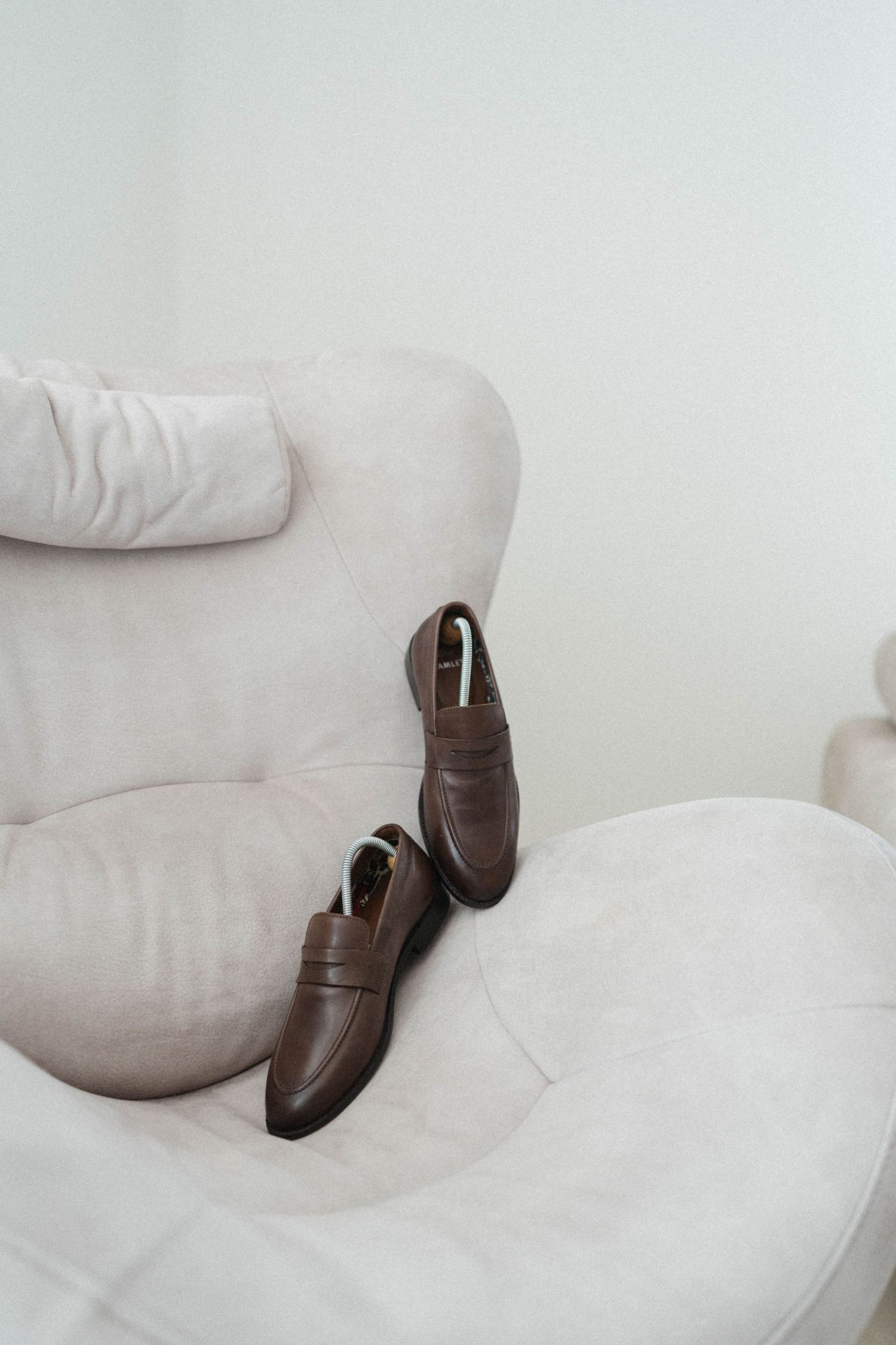 Brown leather penny loafers with shoe trees resting on a cream velvet armchair.