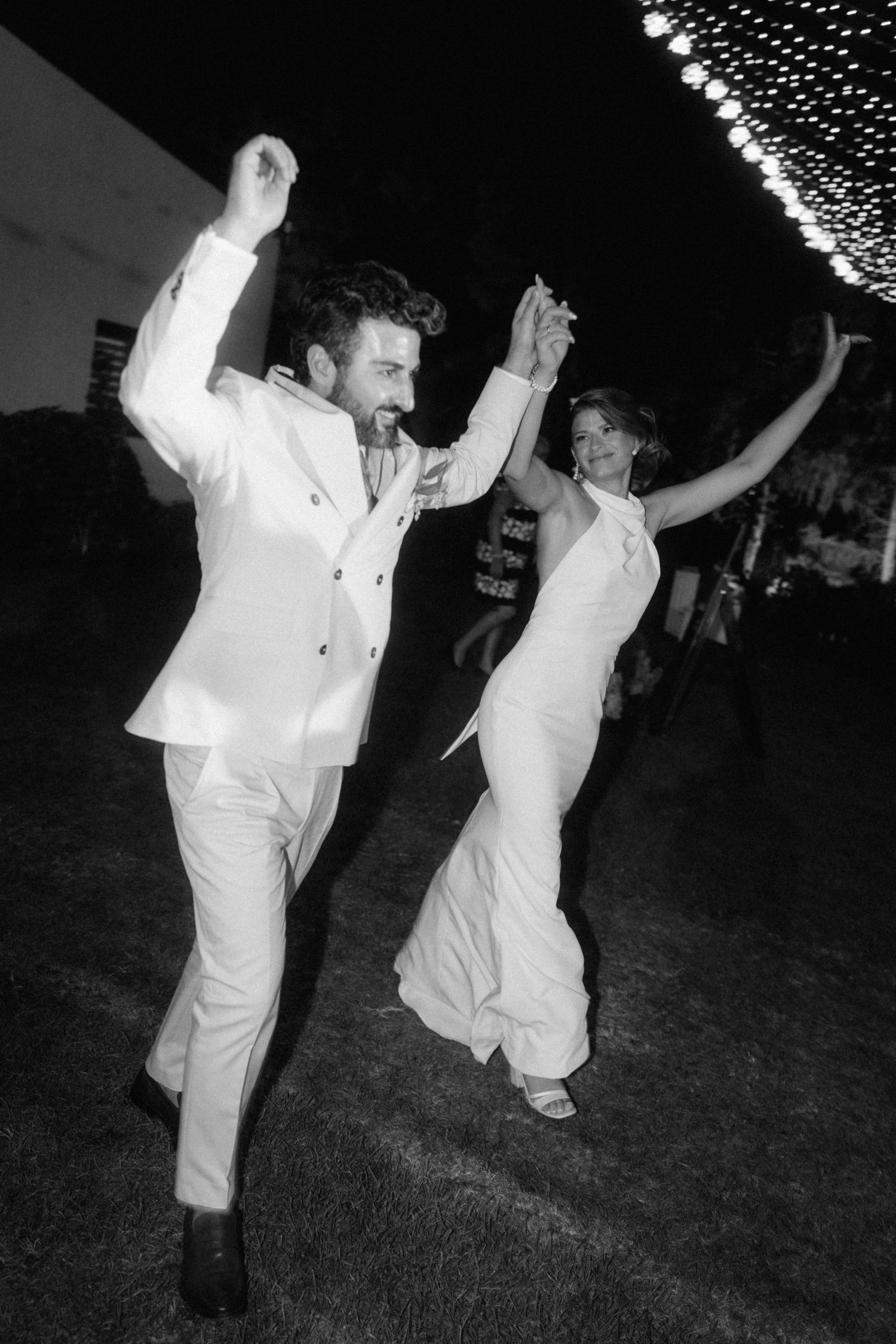 Black and white photo of bride and groom dancing energetically at outdoor wedding reception under string lights.