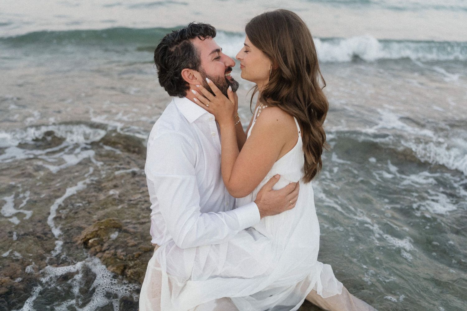Romantic couple in white outfits sharing a tender moment by ocean waves on rocky shore at sunset.