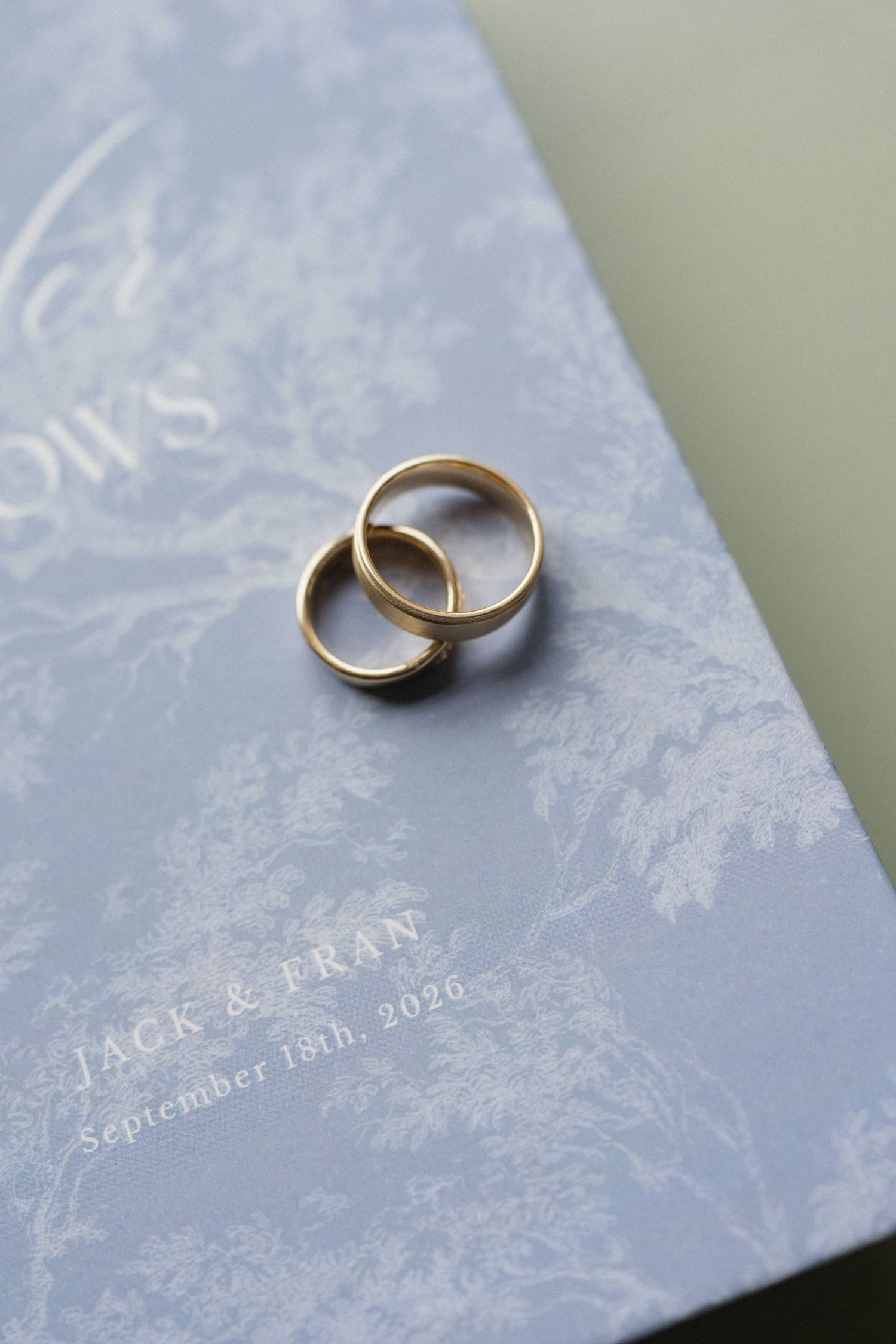 Two gold wedding bands rest on a blue vow book with a toile pattern, printed with Jack & Fran and the date September 18th, 2026.
