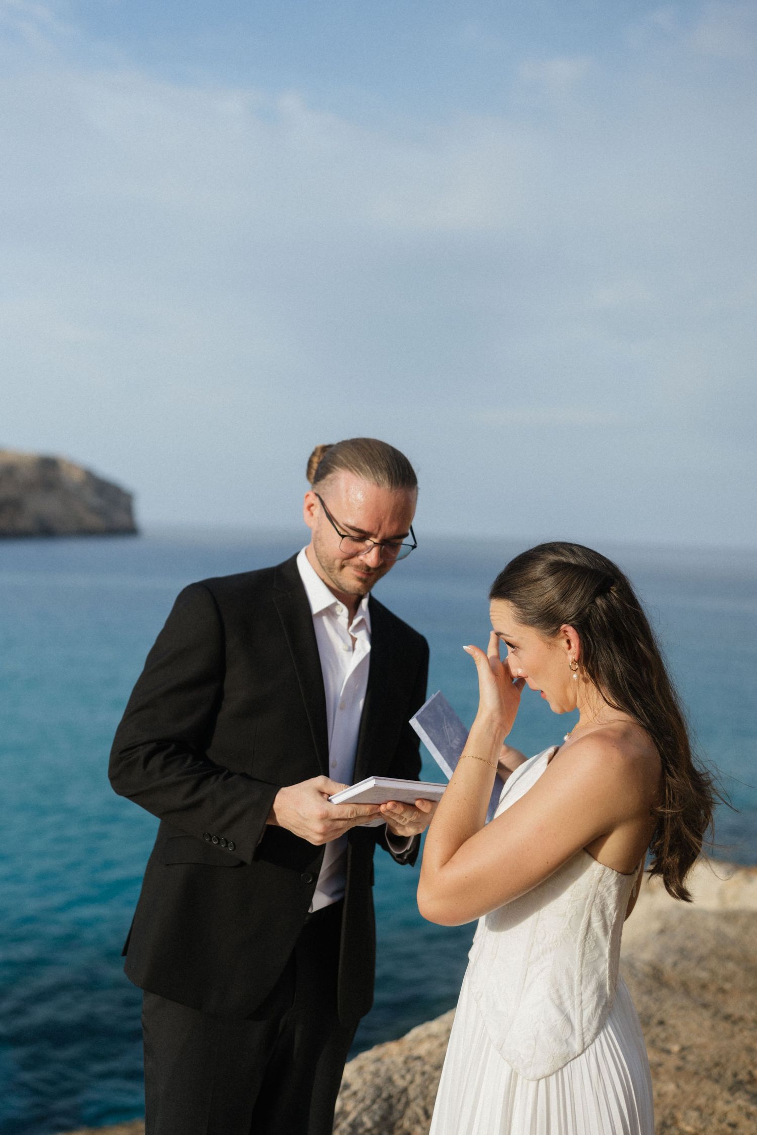 Bride wipes away tears while the groom in glasses and a black suit reads vows from a book during an oceanside cliff wedding ceremony.