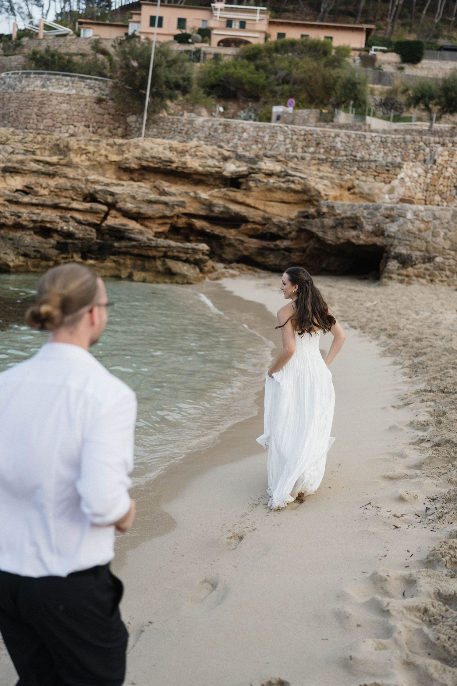 Bride in a white dress walks barefoot along a sandy beach toward rocky cliffs as a man in a white shirt follows beside the calm water.