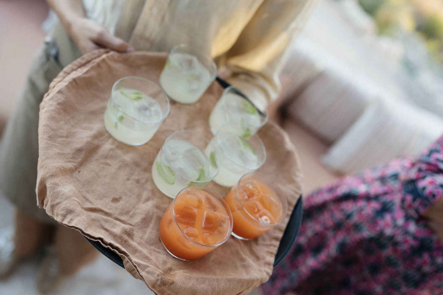 Server carrying a linen-lined tray of refreshing cocktails in glasses with ice and citrus garnish at a wedding reception.