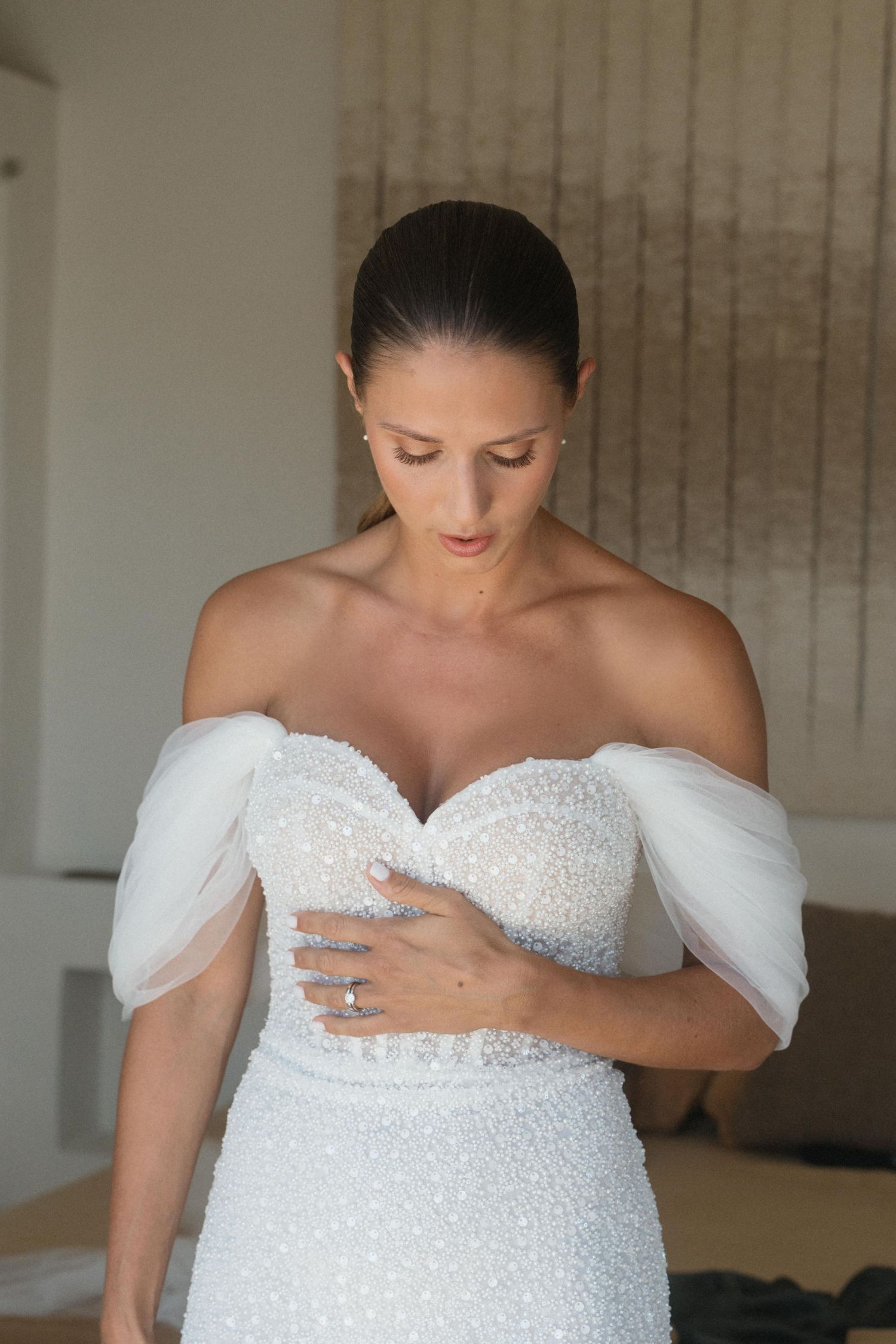 Bride in a beaded off-the-shoulder wedding gown with sheer tulle sleeves looks down while resting her hand on her waist.
