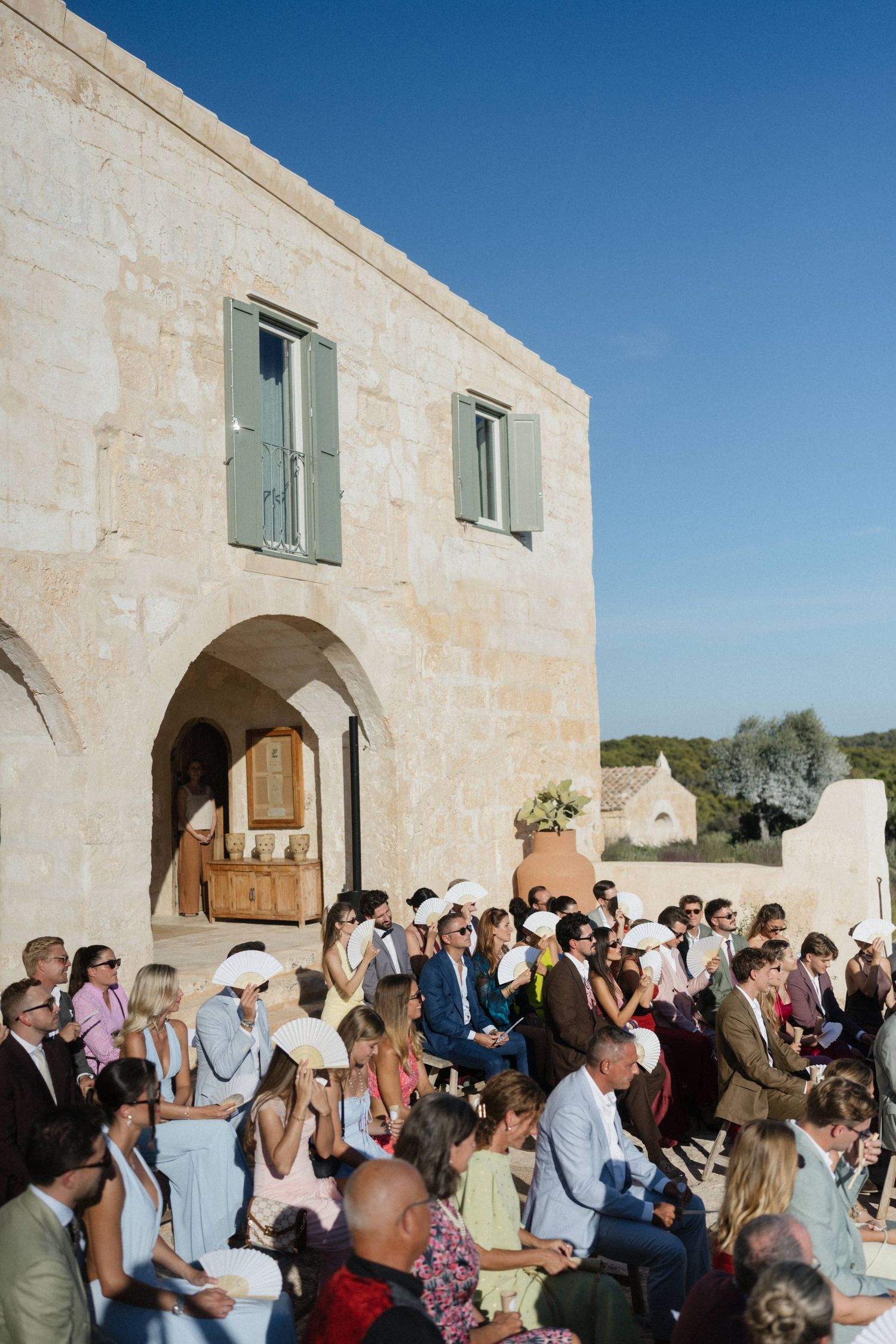 Wedding guests seated outdoors watch a ceremony beside a rustic stone building with green shutters under a clear blue sky.