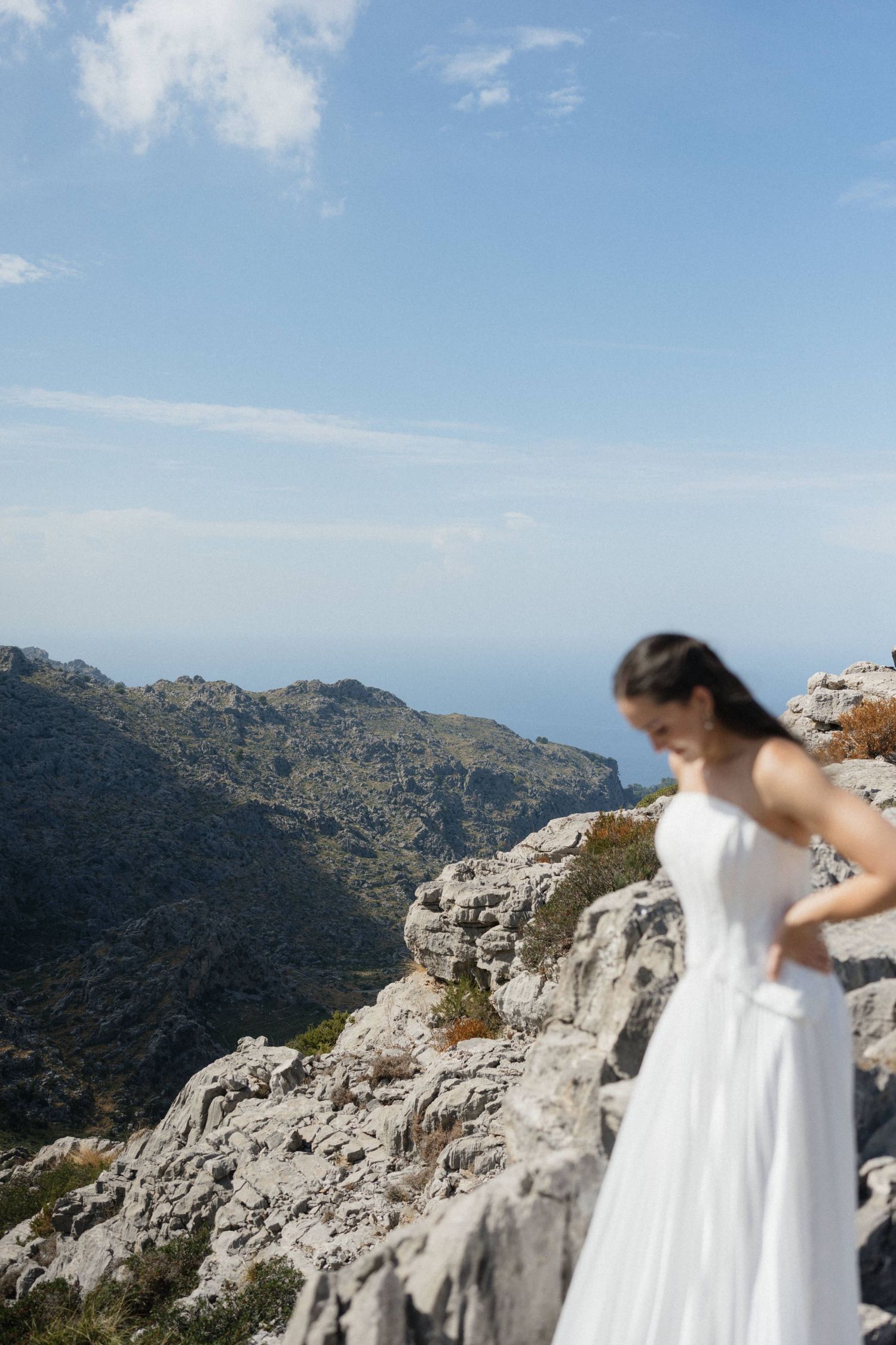 Blurred bride in a flowing white dress stands on a rocky mountain ledge overlooking distant hills and the sea under a blue sky.