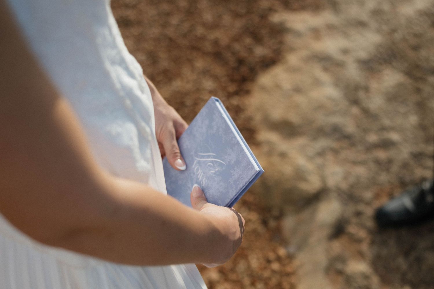 Bride in a white dress holds a small blue vow book while standing on sandy, rocky ground during an outdoor elopement ceremony.