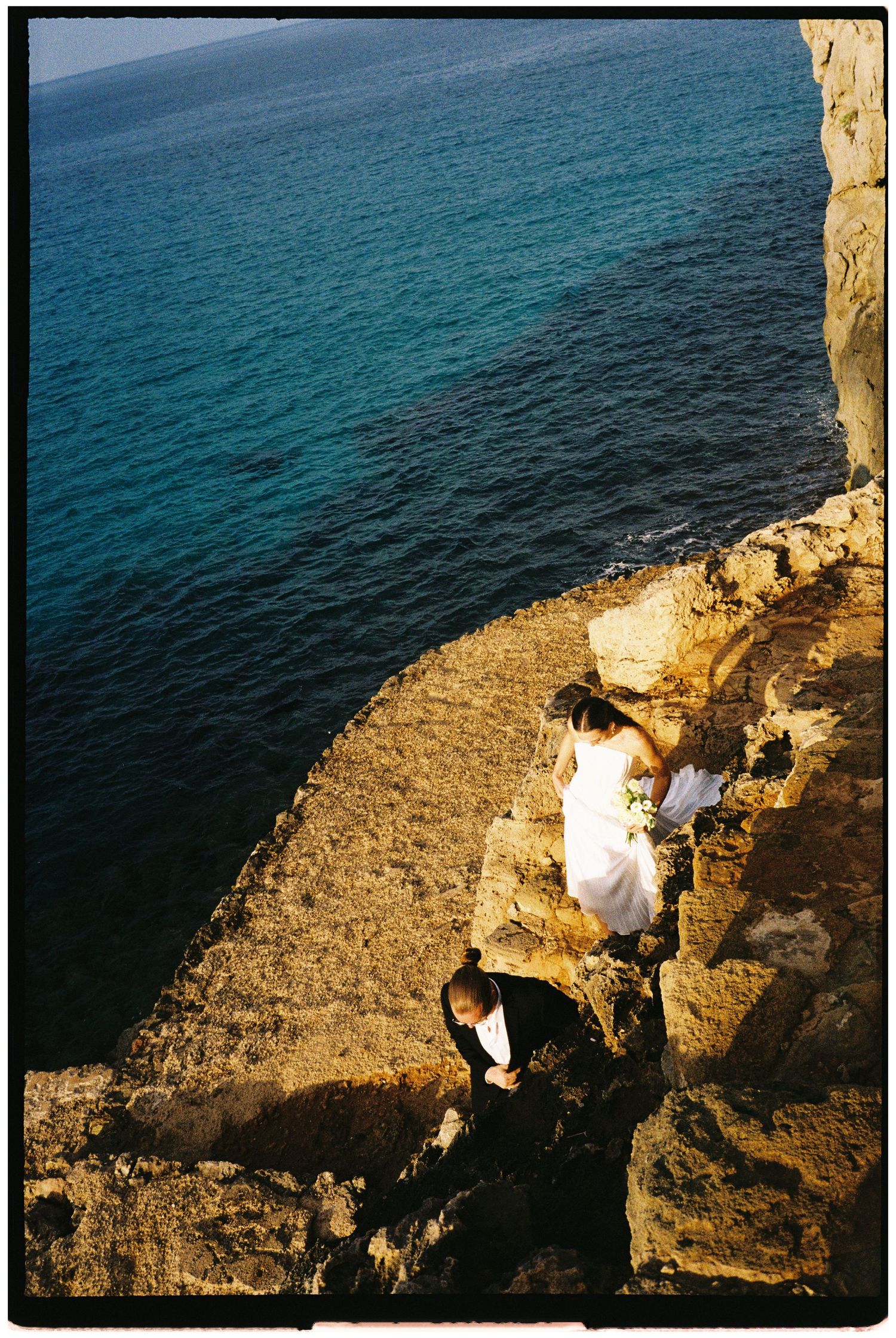 Bride in a white gown and groom in a dark suit climb rocky seaside cliffs above the turquoise Mediterranean for a scenic wedding photo.