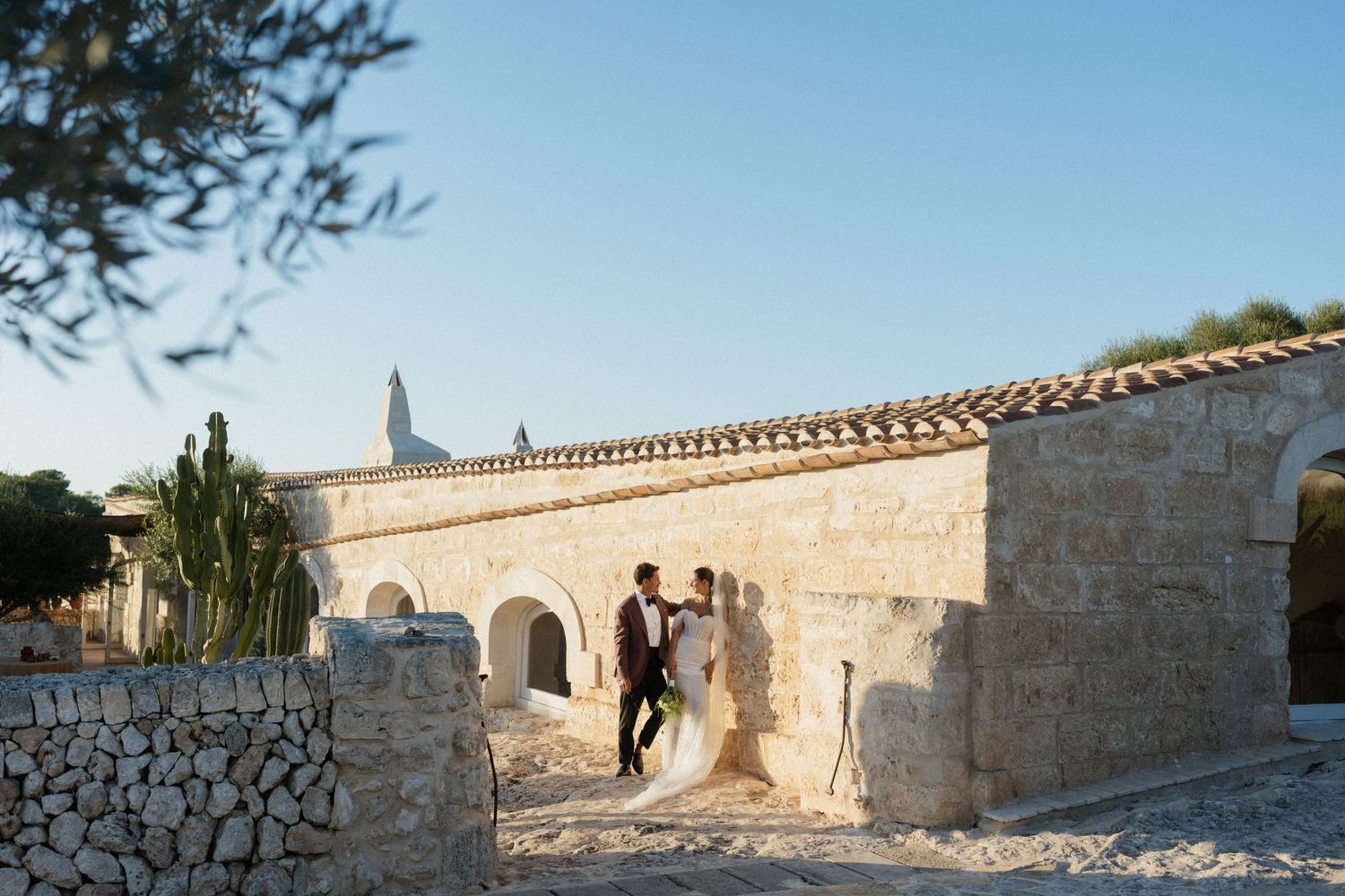 Bride and groom embrace beside a rustic stone building with arched doorways and a bell tower under a clear blue sky.