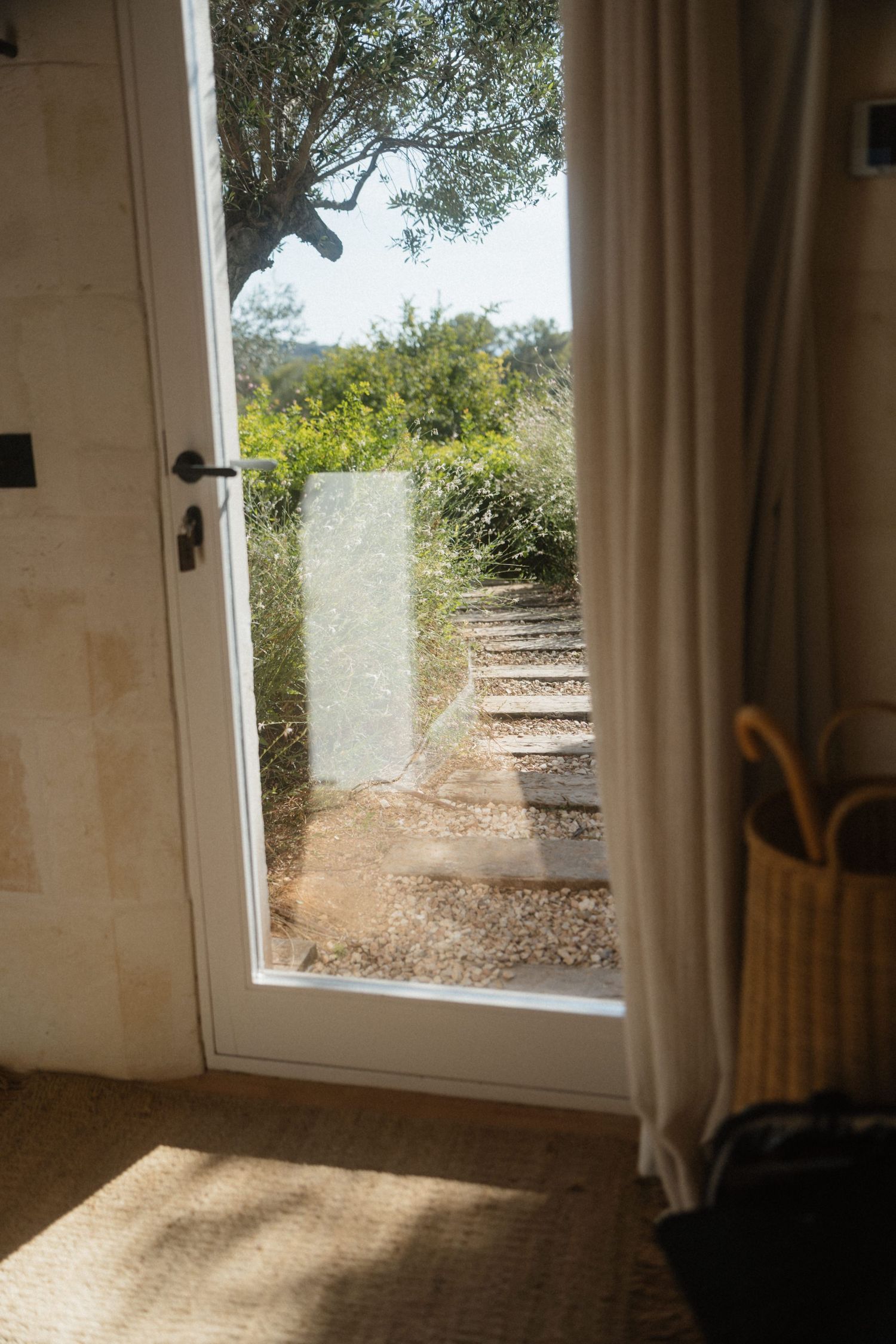 Sunlit glass door opening onto a stone path through olive trees and greenery, with a woven basket and linen curtain nearby.