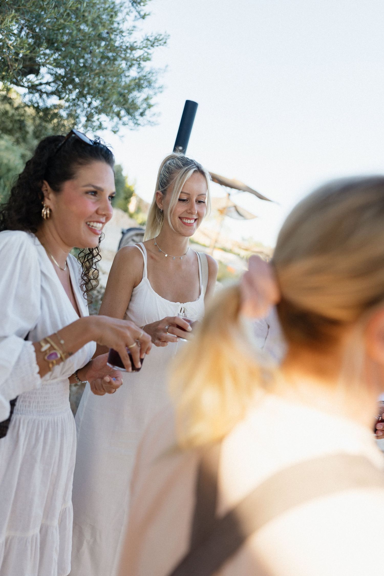 Smiling women in white summer dresses chat and laugh together at a sunny outdoor garden party.