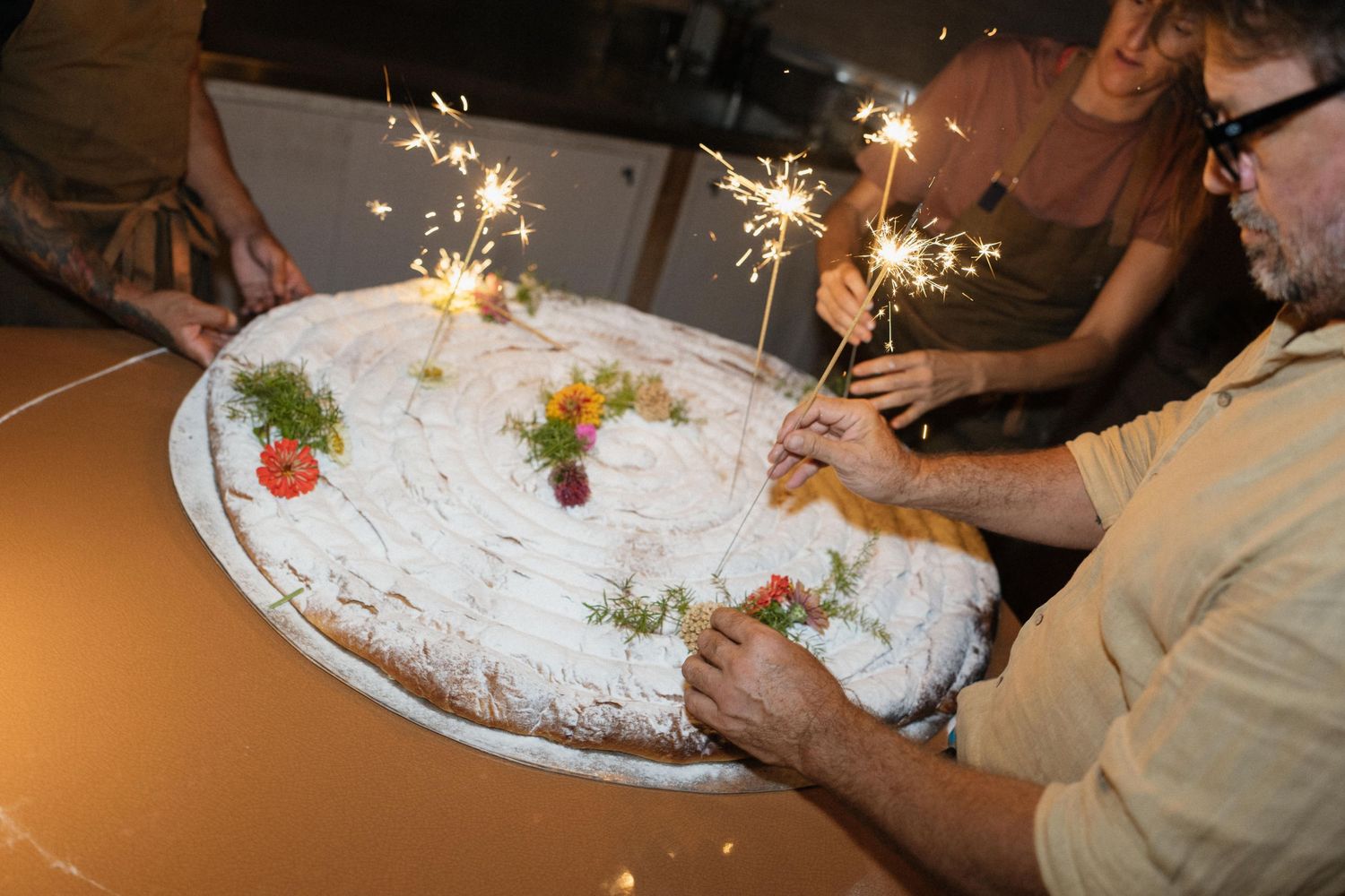Guests gather around a large round cake decorated with flowers and lit sparklers, celebrating at a festive table.