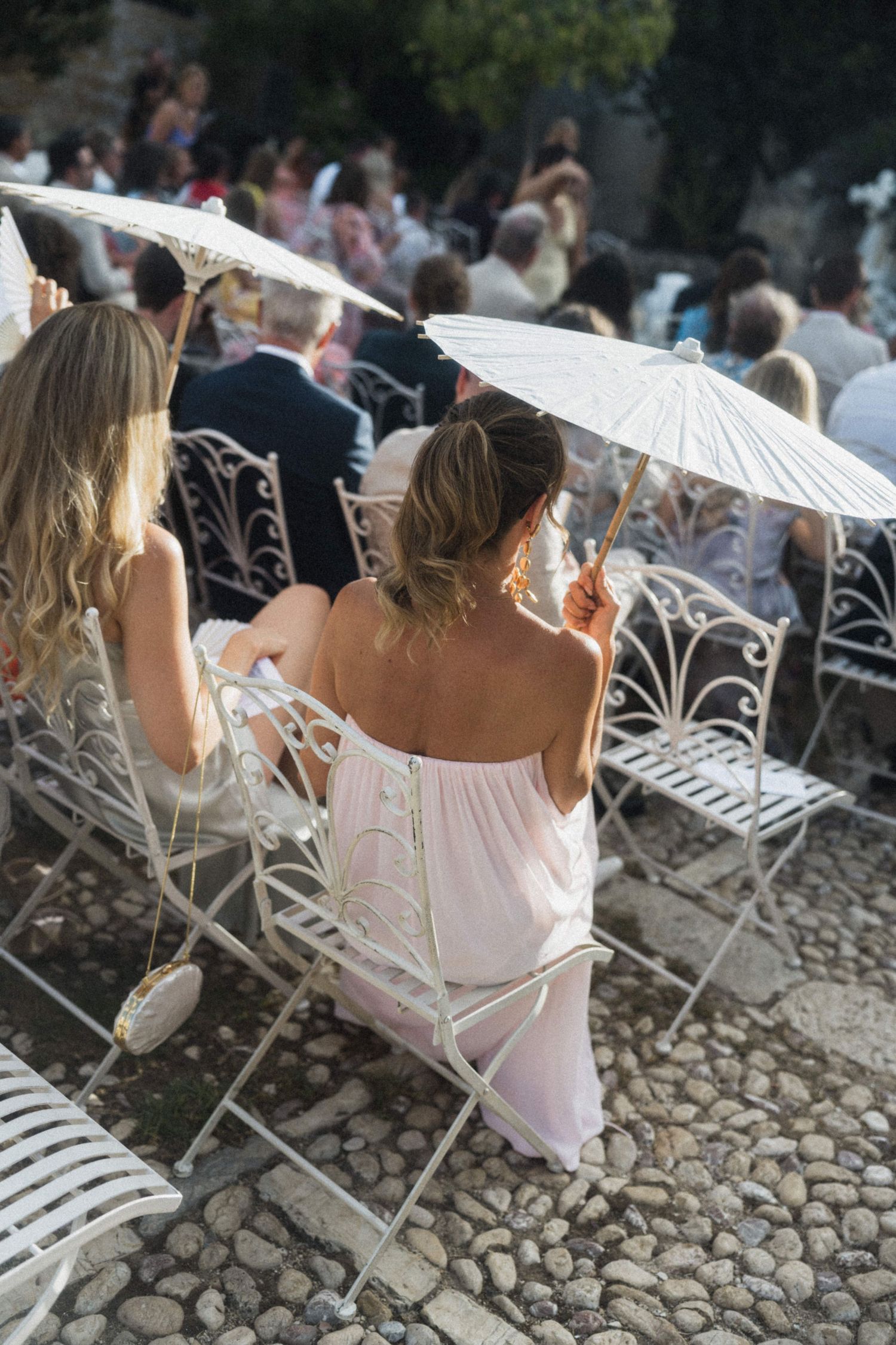 Wedding guests seated in ornate white chairs holding parasols at outdoor ceremony.