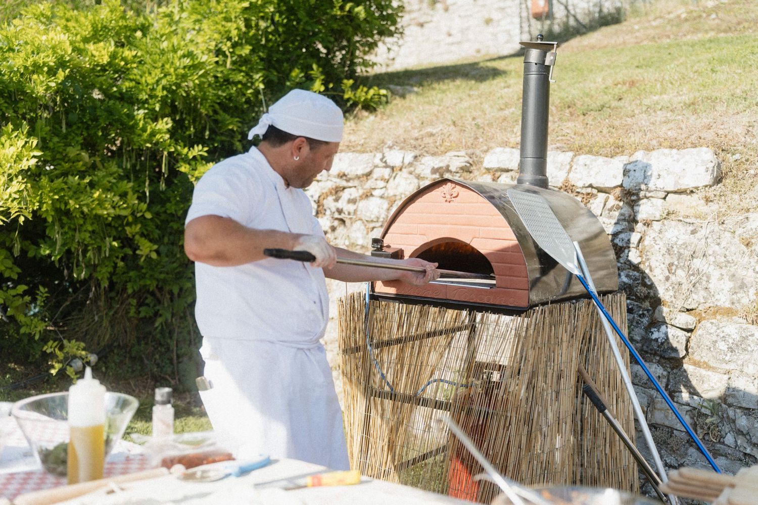 Chef in white uniform placing a pizza into a traditional wood-fired oven at an outdoor event.