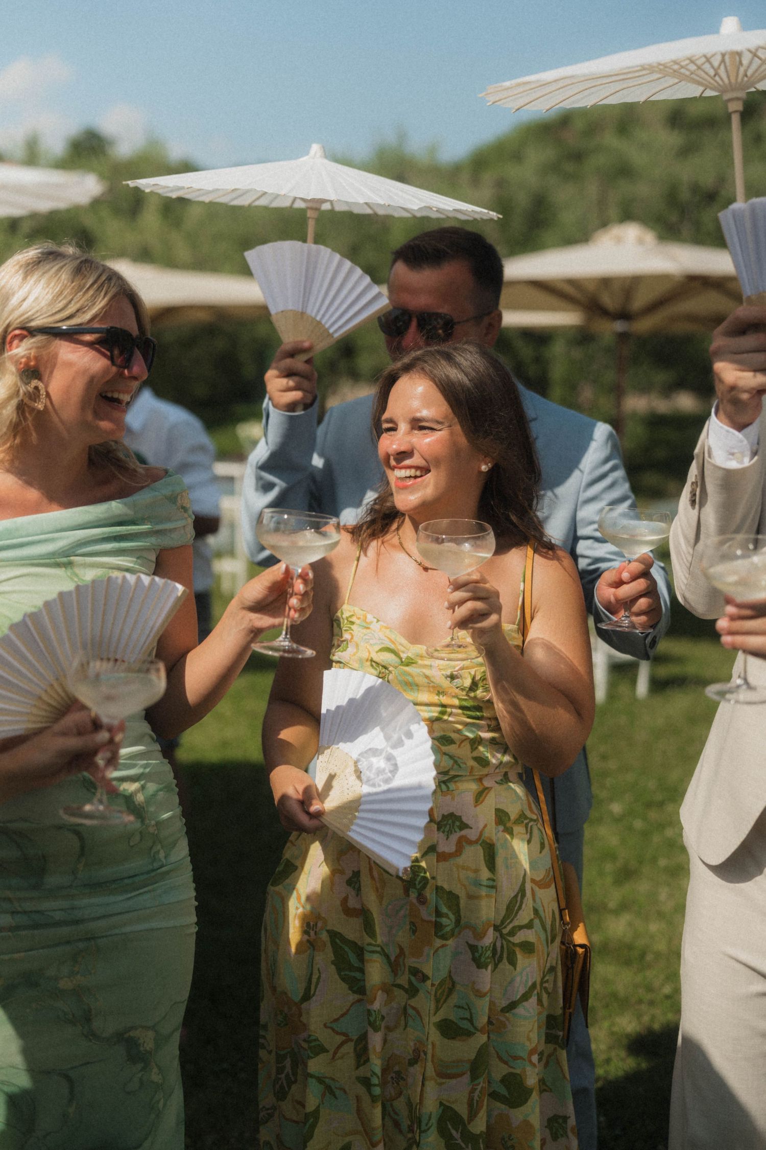 Wedding guests laughing and toasting with cocktails while holding fans and parasols outdoors.