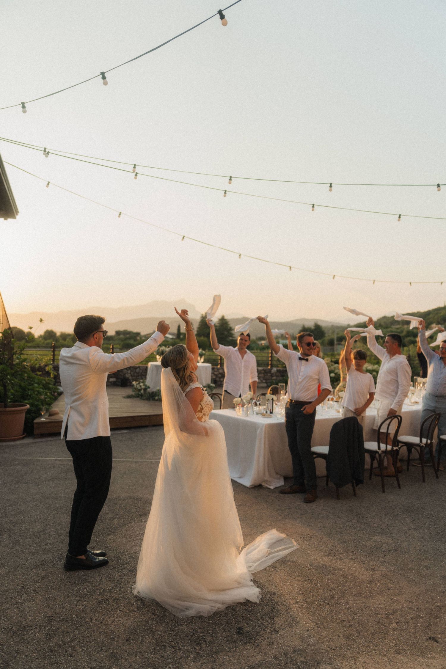 Bride and groom celebrate with guests waving napkins at sunset outdoor wedding reception.
