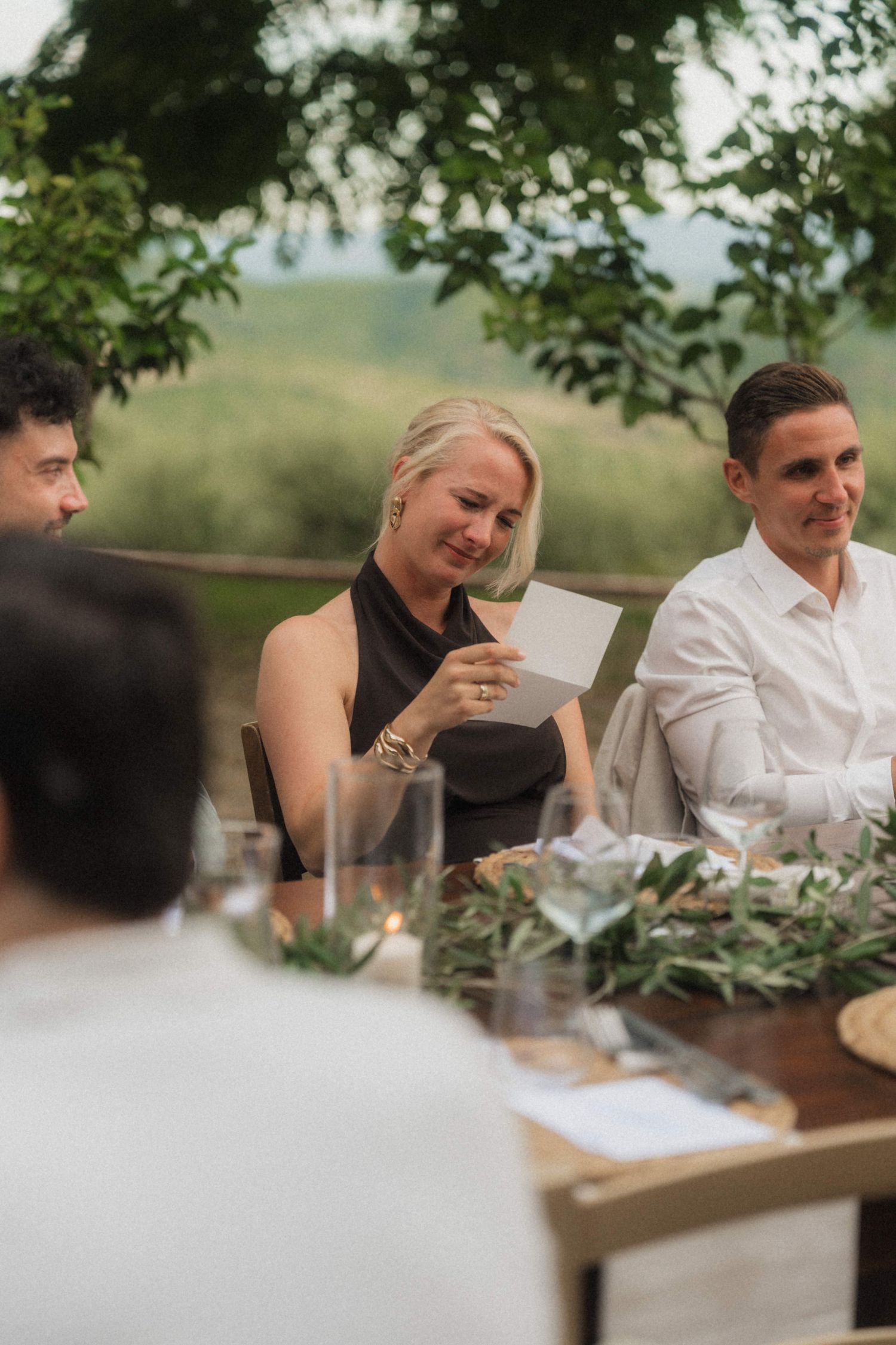 Wedding guest in black dress reading card at elegant outdoor reception dinner table.