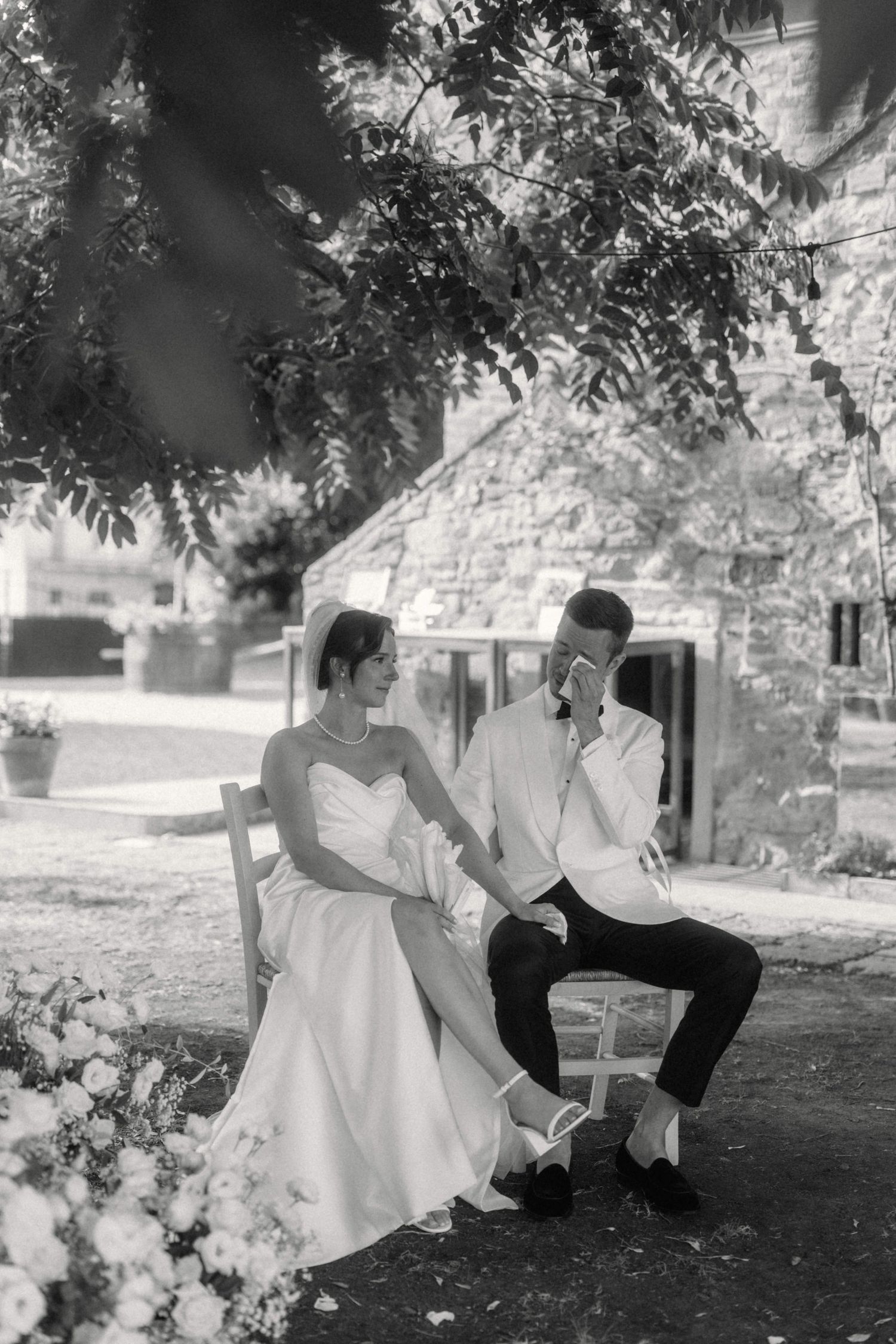 Black and white photo of bride and groom sitting together on chairs beneath a tree at their wedding venue.
