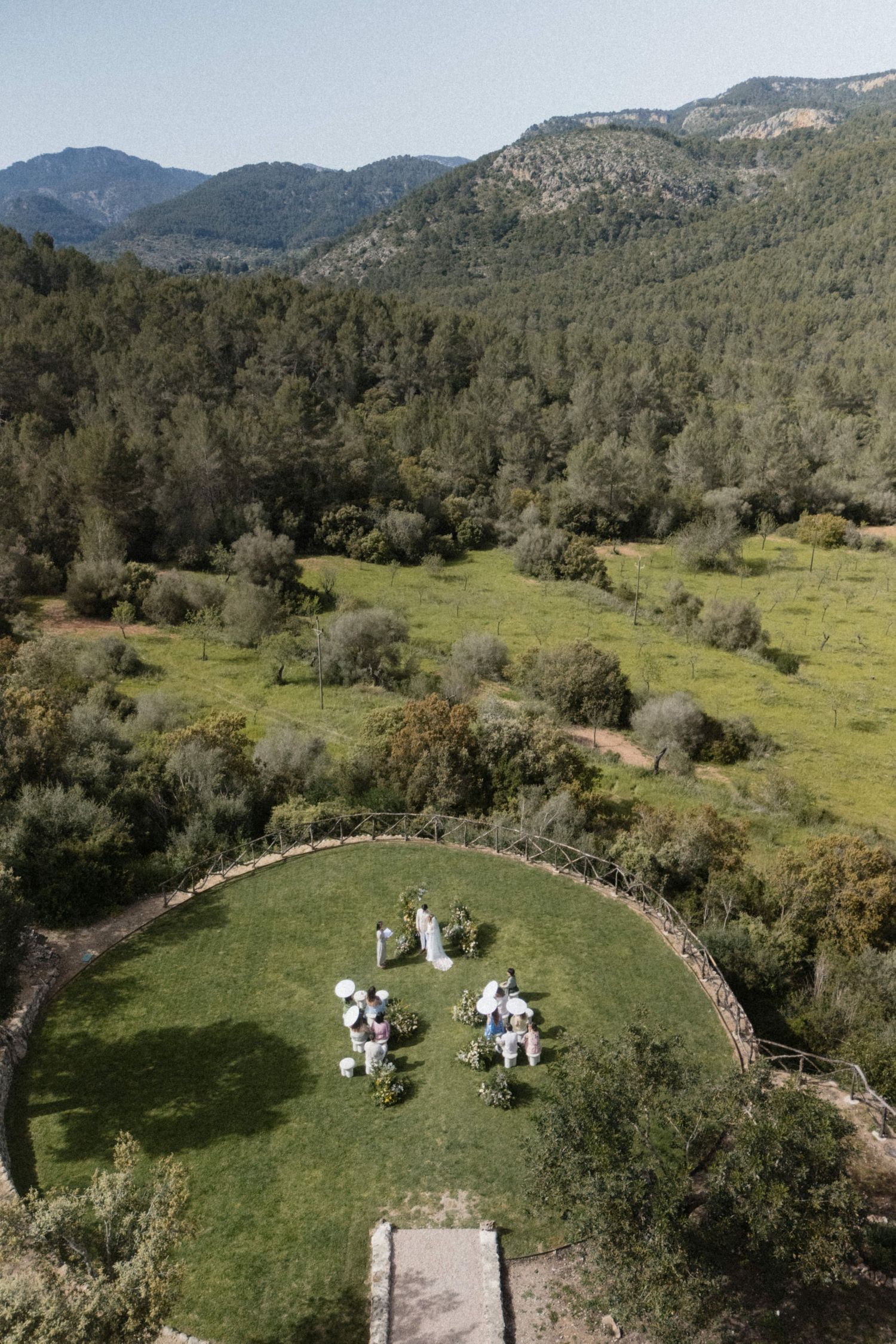 Drone aerial view of an intimate outdoor wedding ceremony on a circular lawn with stunning mountain scenery.