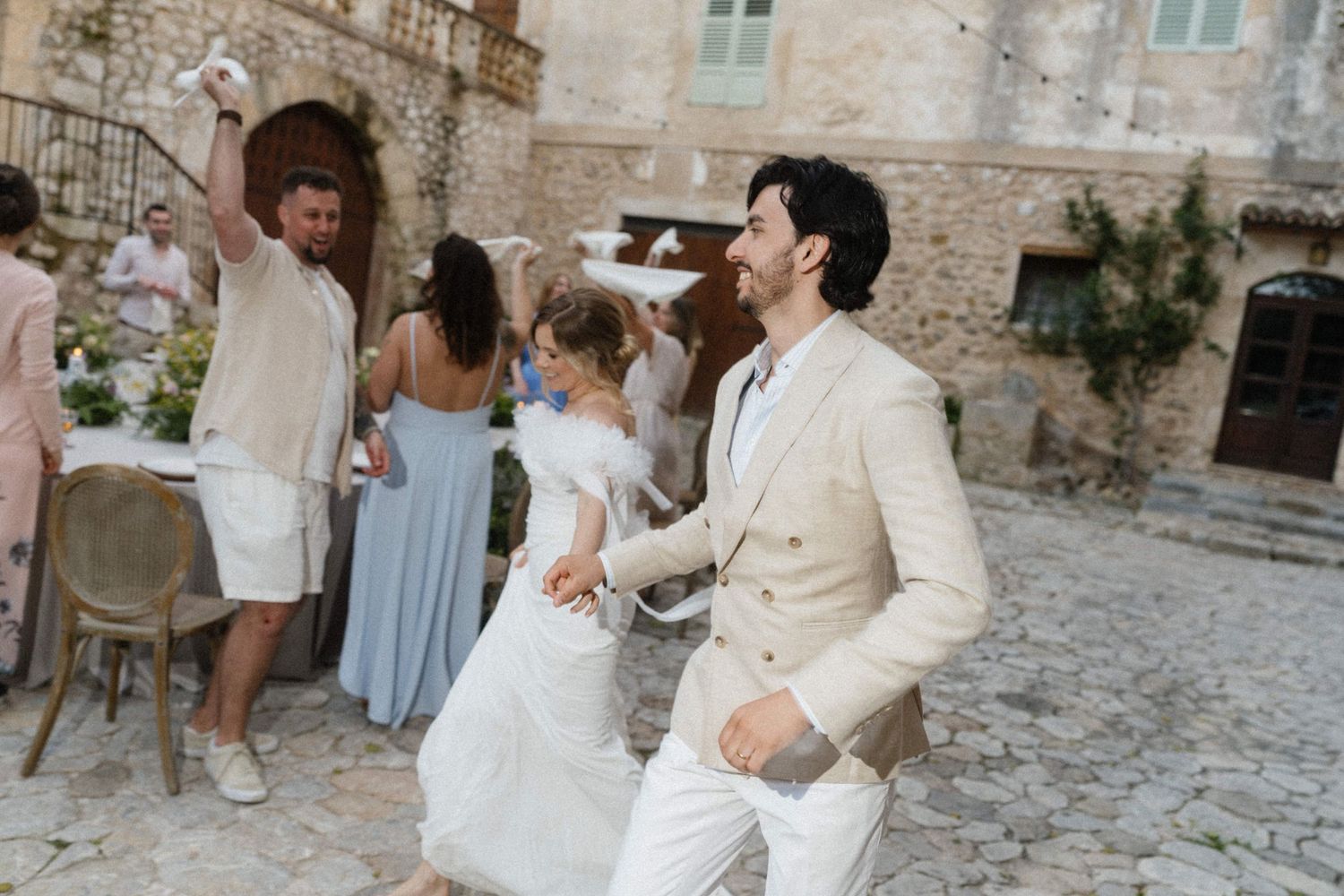 Bride and groom dancing joyfully in a rustic stone courtyard surrounded by wedding guests.