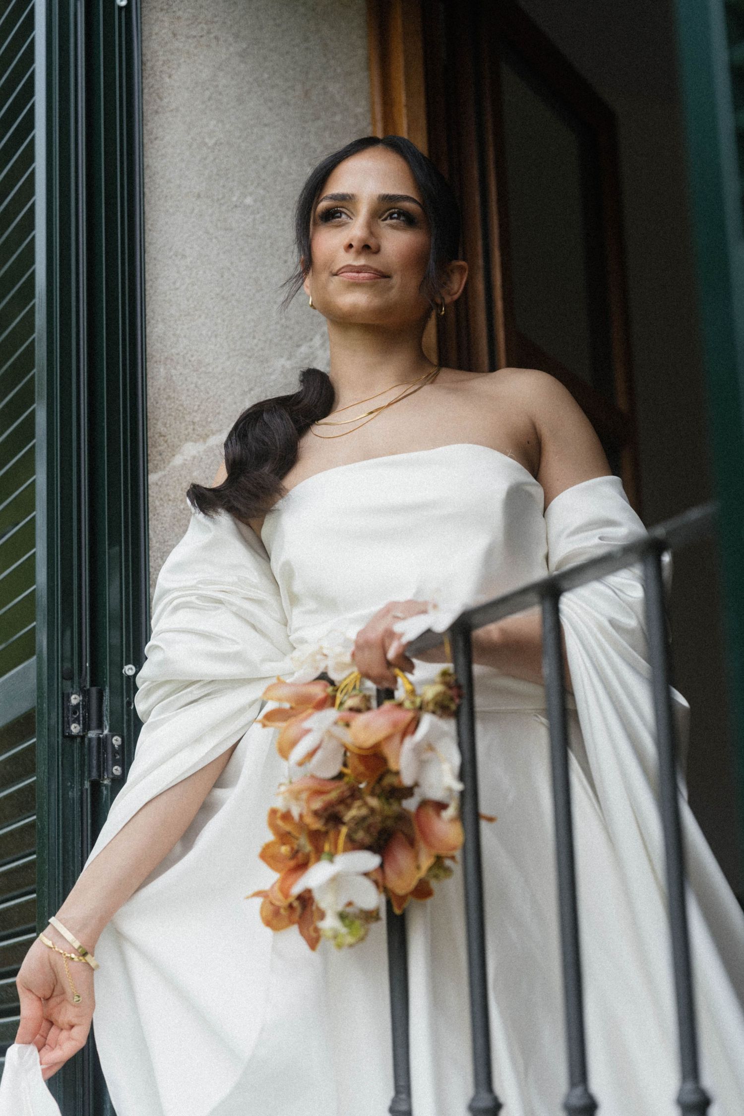 Beautiful bride in strapless white gown holding autumn floral bouquet on balcony of stone building.