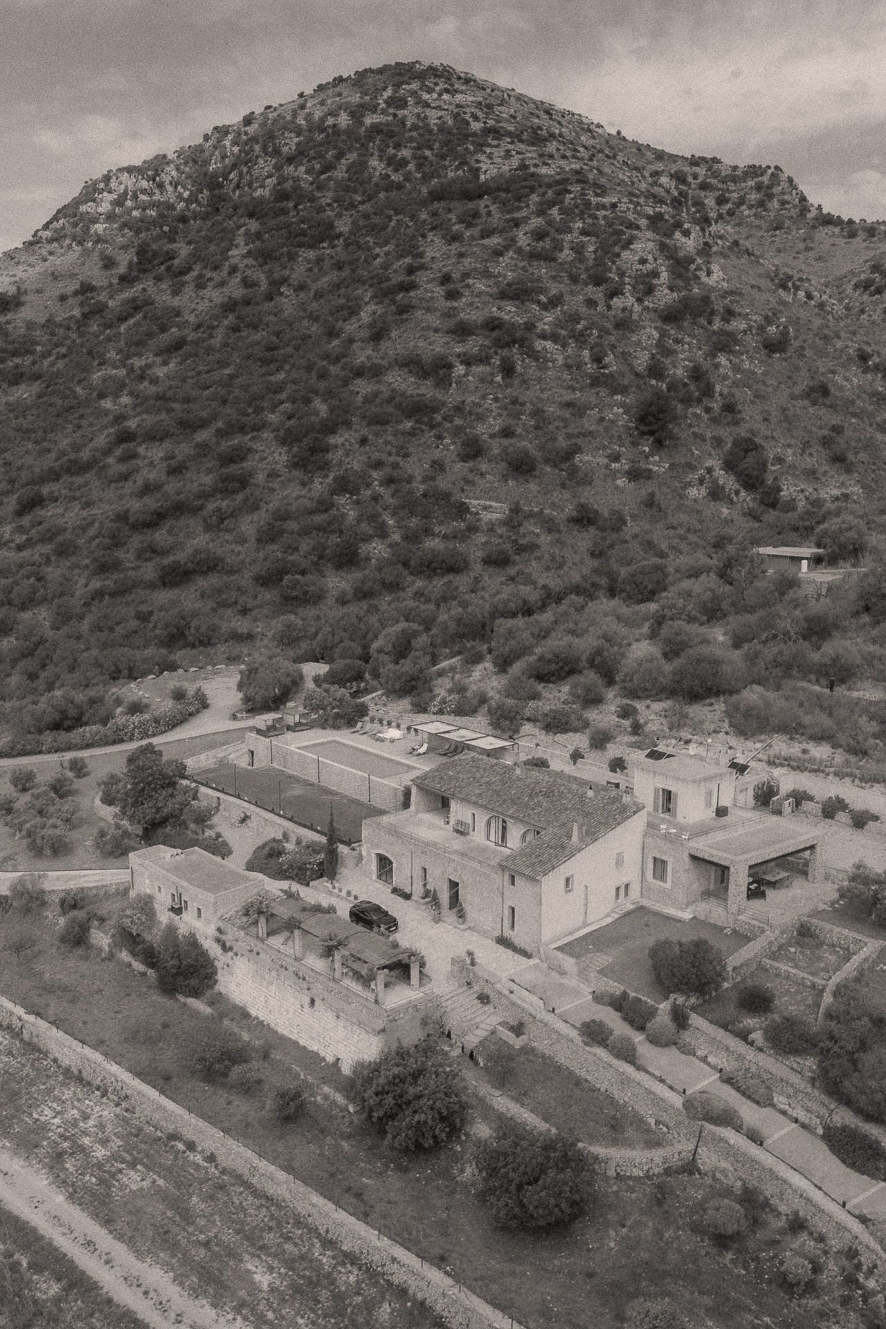 Aerial black and white view of a rustic Mediterranean estate nestled at the foot of a lush tree-covered mountain.