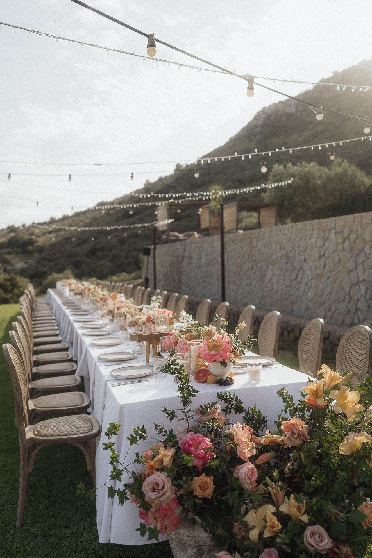 Elegant outdoor wedding reception table with pink floral arrangements, string lights, and white linens set against a stone wall.