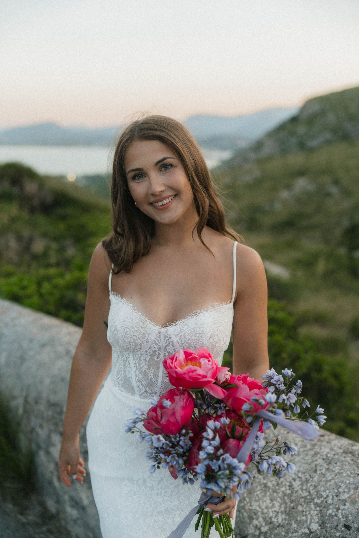 Smiling bride in a white lace gown holding a vibrant pink peony bouquet with scenic mountain backdrop.