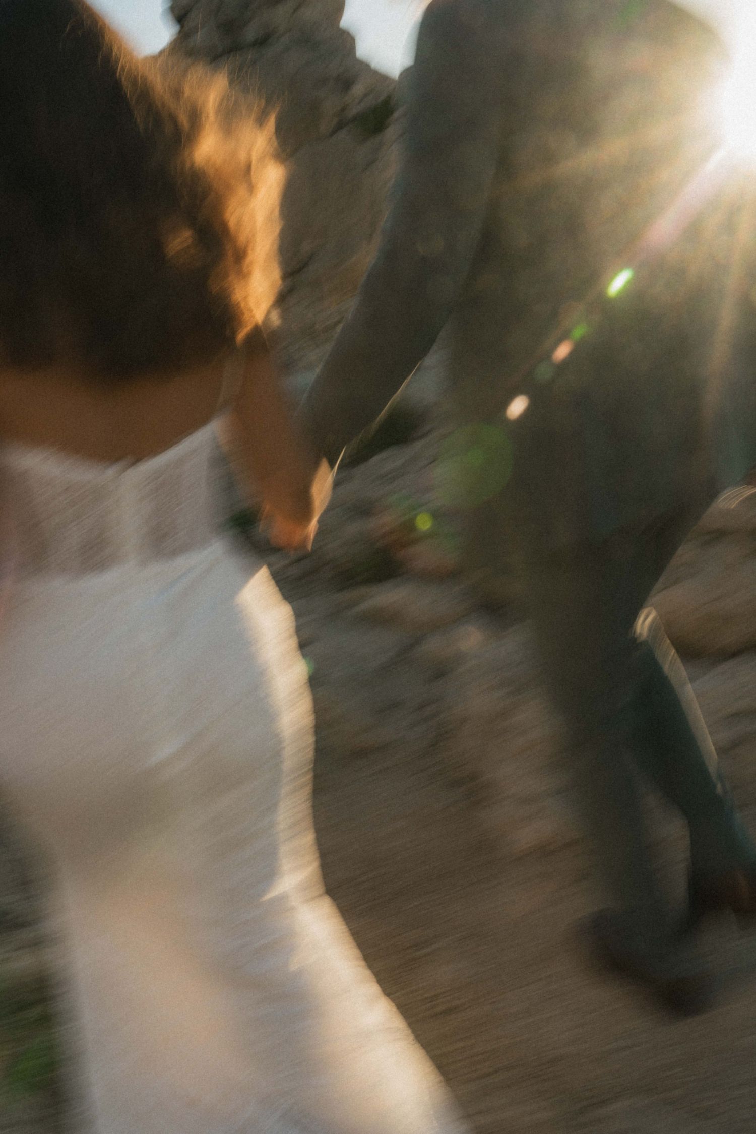 Blurred motion shot of a bride and groom holding hands, walking among rocks with a golden sun flare behind them.